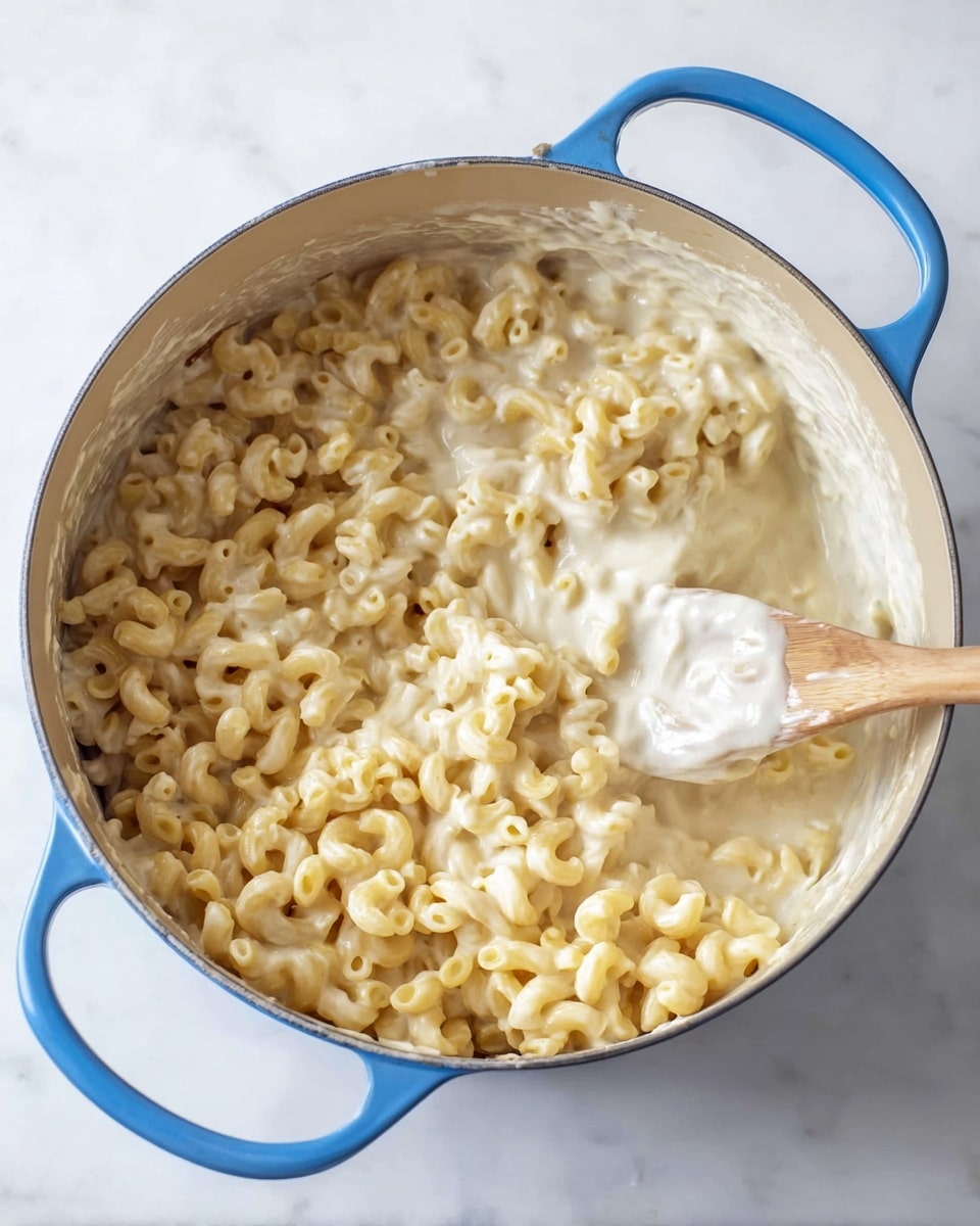 A blue pot filled with creamy white cheese sauce and pale yellow elbow macaroni pasta being stirred with a wooden spoon, showing a mix of smooth, thick sauce on one side and pasta on the other. The pot sits on a white marbled surface. photo taken with an iphone --ar 4:5 --v 7