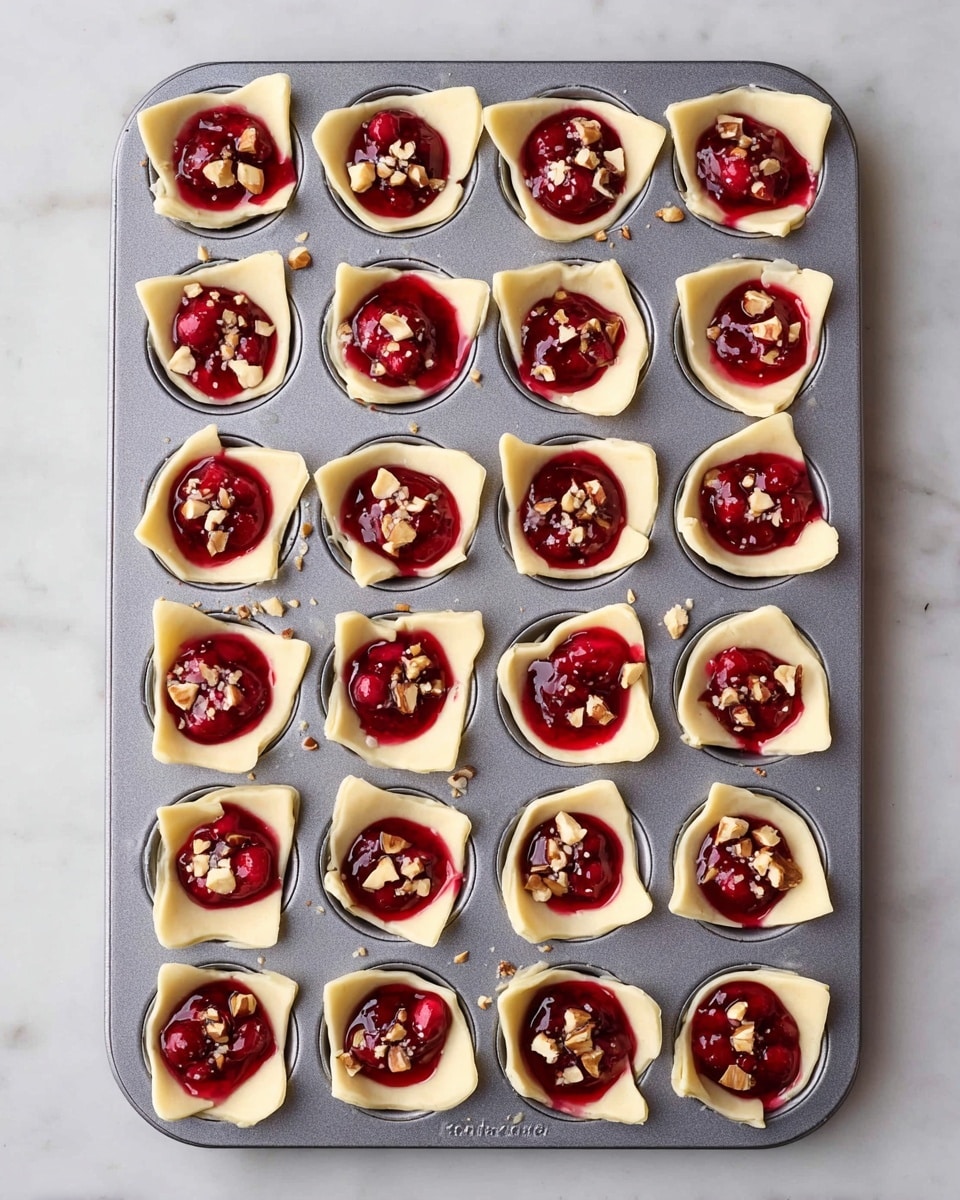 Twelve small puff pastry squares are arranged on a beige plate with each square having a golden, flaky base layer that is puffed up around the edges. In the center of each pastry is a vibrant red layer of cranberry sauce, topped with bits of melted pale yellow cheese, small pieces of chopped nuts, and a fresh green rosemary sprig. The plate sits on a white marbled surface with some loose cranberries and rosemary sprigs scattered nearby. Photo taken with an iphone --ar 4:5 --v 7