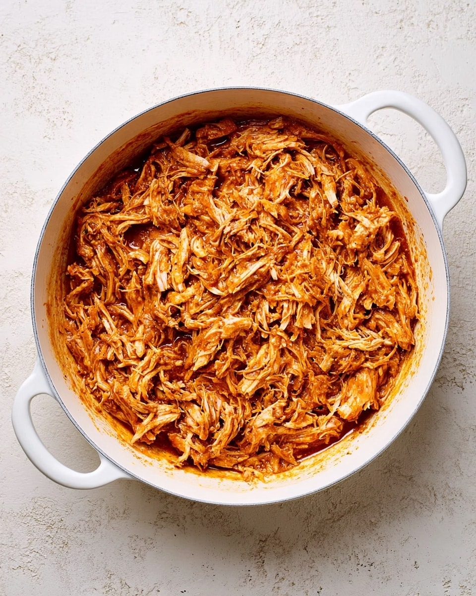 The image shows a top view of a white round pan with two side handles, filled with shredded orange-brown meat that looks tender and juicy. The meat is mixed with a thick sauce that covers every piece, giving it a slightly shiny texture. The surface around the pan is a white marbled texture, clean and simple, making the pan and meat stand out. Photo taken with an iphone --ar 4:5 --v 7