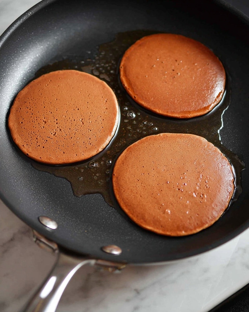 The image shows three round brown pancake batter circles cooking on a black non-stick pan placed on a stove. The pancakes have a smooth, shiny surface with a few small bubbles appearing on top, indicating they are just starting to cook. The pan has some oil glistening around the pancakes, giving a moist look to the cooking surface. The pan’s silver handle and rivets are slightly visible on the left side, and the background shows a white marbled texture. photo taken with an iphone --ar 4:5 --v 7