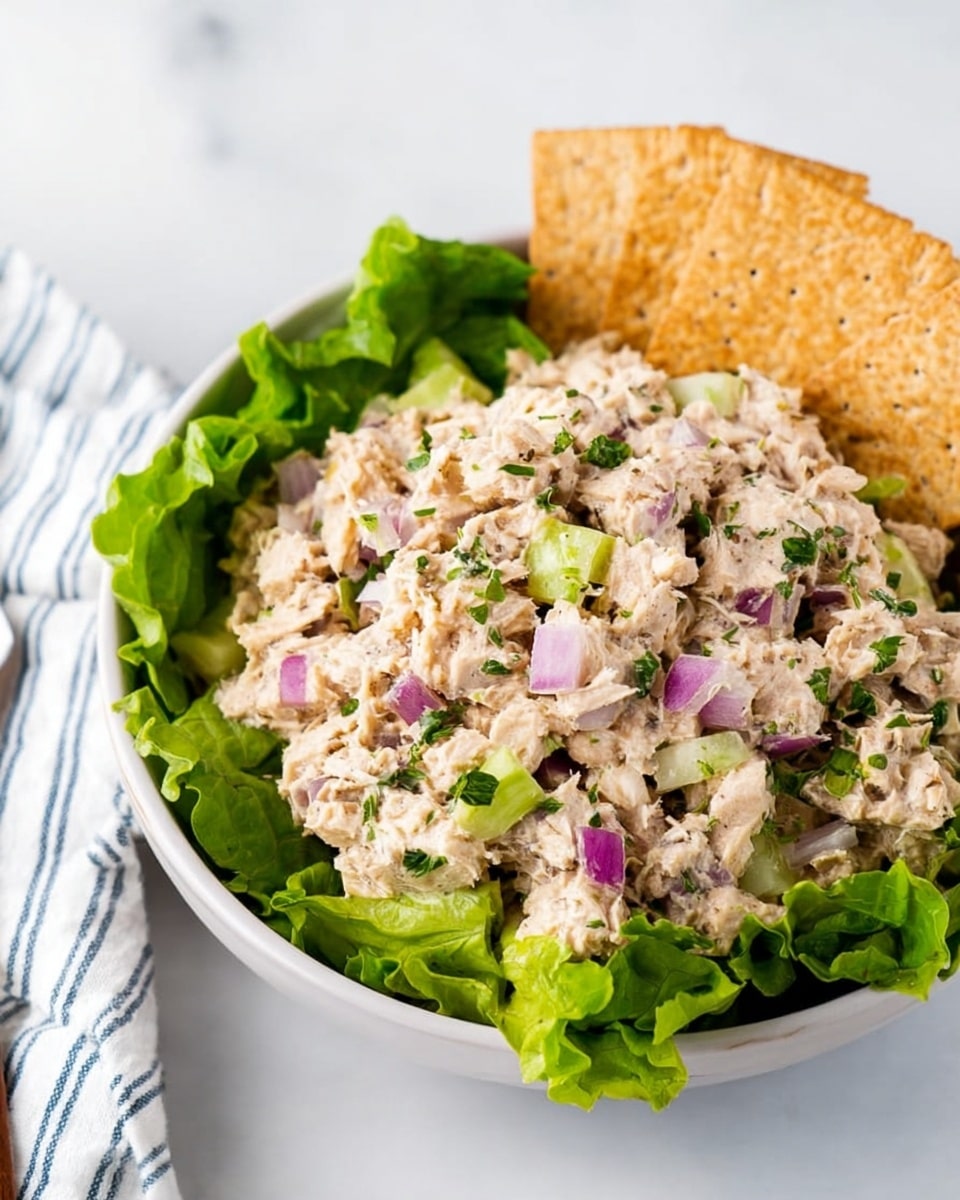 A white bowl contains a mixed tuna salad on a bed of green leaf lettuce. The salad has small pieces of light beige tuna mixed with diced bright purple onions and light green celery. There are green herb sprinkles on top, adding texture and color. On the right side of the bowl, three light brown seeded crackers stand upright against the salad. The bowl sits on a white marbled surface with a white cloth featuring blue stripes nearby photo taken with an iphone --ar 4:5 --v 7
