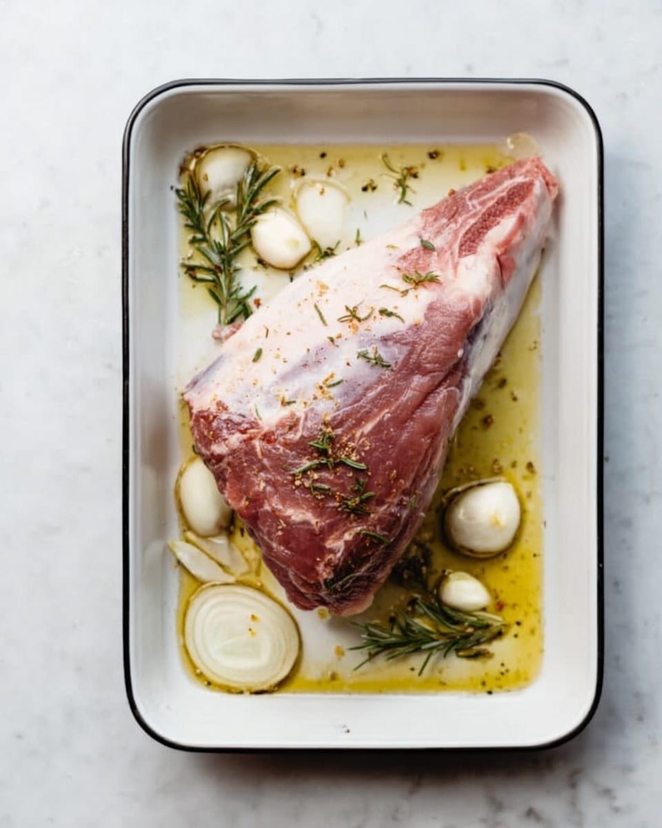 A single piece of raw meat, light pink and marbled with fat, lies in a rectangular white baking tray filled with clear olive oil. Around the meat, there are onion slices and whole peeled garlic cloves with their skin, scattered evenly. Small sprigs of fresh rosemary are also placed around the meat, adding green accents to the scene. The tray sits on a white marbled surface. photo taken with an iphone --ar 4:5 --v 7