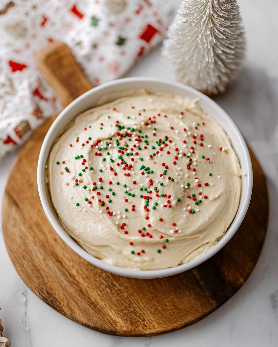 A round white bowl filled with one smooth layer of light beige creamy mousse or frosting, evenly spread with soft swirling texture on top. The surface is sprinkled with small red, green, and white round sprinkles scattered evenly. The bowl sits on a wooden board, and around it, there is a blurred white marbled texture background with holiday-themed paper and a white frosted small tree decoration visible. photo taken with an iphone --ar 4:5 --v 7