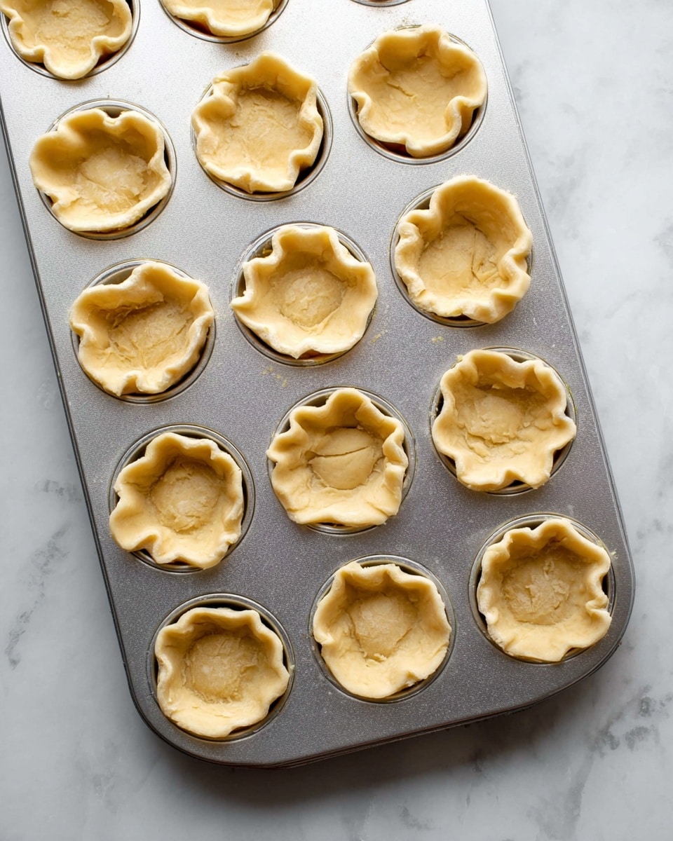 A metal muffin tray holds fifteen small pie crusts, each shaped roughly with ruffled edges pressed into the tray's round wells. The dough is light golden with a soft texture, some crusts having uneven folds and creases, giving a homemade look. The tray rests on a white marbled surface, adding a clean and bright background to the simple, unbaked pastry shells photo taken with an iphone --ar 4:5 --v 7