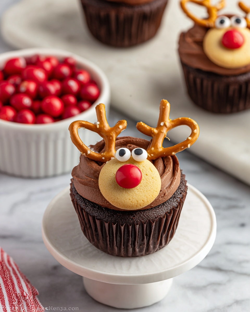 A chocolate cupcake sits on a small white pedestal with three layers: the dark brown cupcake base, a thick layer of smooth chocolate frosting covering the top, and a large round golden cookie placed near the front center as the reindeer's snout. On the cookie, a single shiny red candy acts as the nose. Above the cookie, two small round white candies with black dots create the eyes. Curving around the top edge of the cupcake are two light brown pretzels forming the antlers. In the background, two similar cupcakes appear on a white marbled surface along with a white bowl filled with red candies and a red and white striped cloth in the corner. photo taken with an iphone --ar 4:5 --v 7