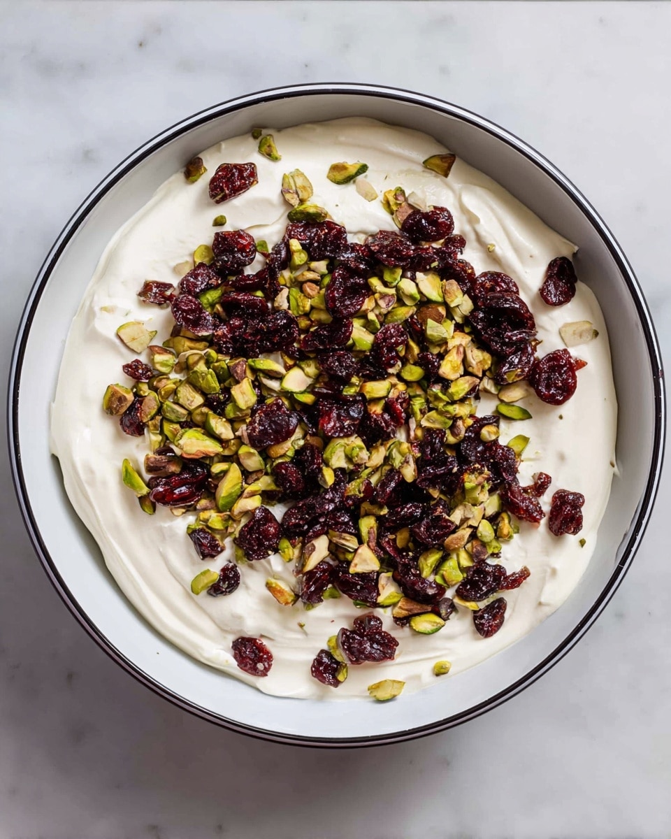 A white plate holds about ten rectangular bars of white chocolate layered with chopped green pistachios and bright red dried cranberries scattered on top and inside the bars. The white chocolate is smooth and shiny, creating a creamy base for the textured bits of nuts and fruit that add pops of color. Nearby, two small white dishes hold more chopped pistachios and whole dried cranberries, set on a white marbled surface. There is a green cloth napkin with fringed edges near the plate. Photo taken with an iphone --ar 4:5 --v 7