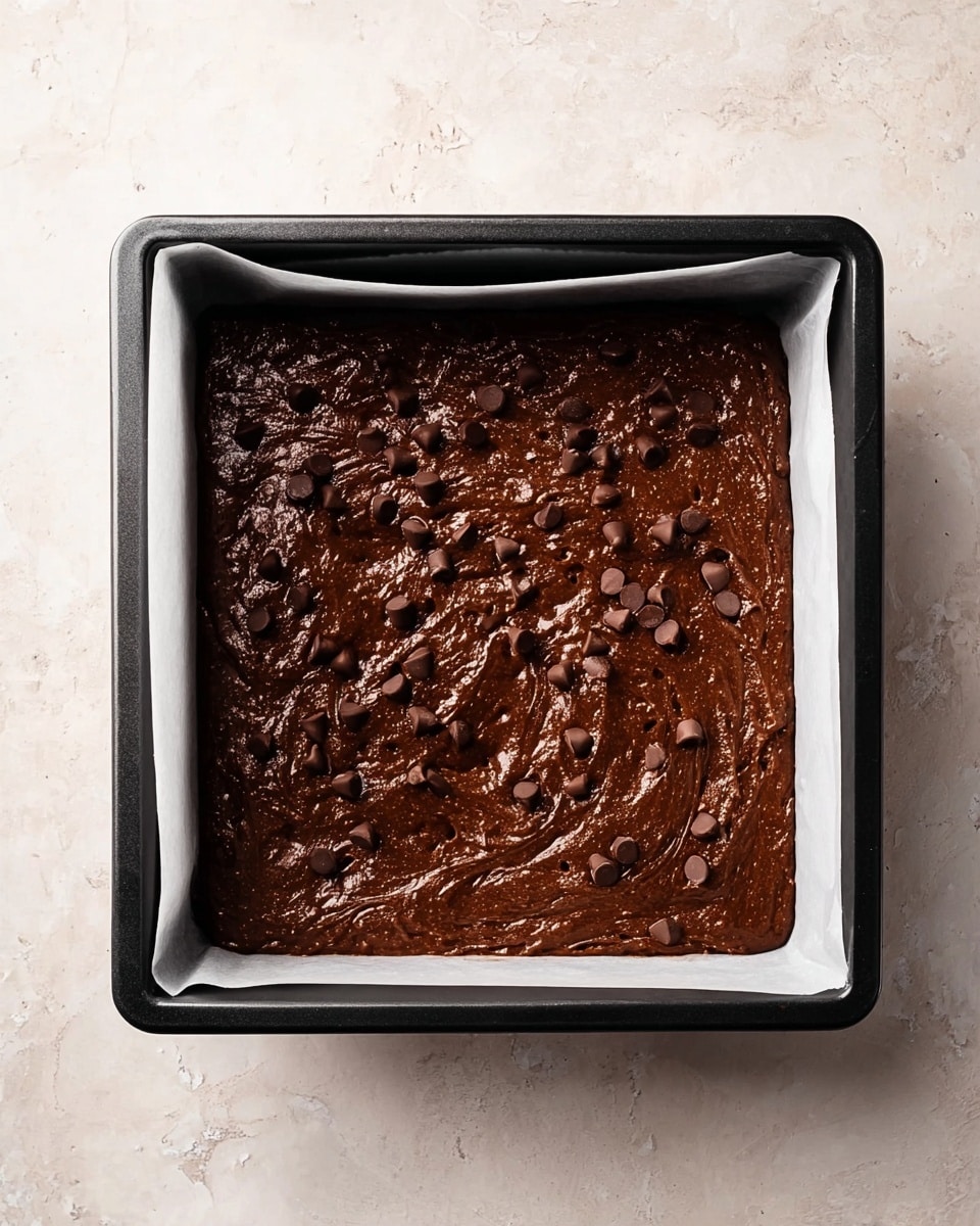 A square black baking tray lined with white parchment paper contains a thick, dark brown chocolate batter spread evenly inside, topped with scattered chocolate chips that add texture and slight shine to the surface. The batter looks smooth and rich, with small swirls and lumps visible throughout. The baking tray rests on a white marbled texture. Photo taken with an iphone --ar 4:5 --v 7