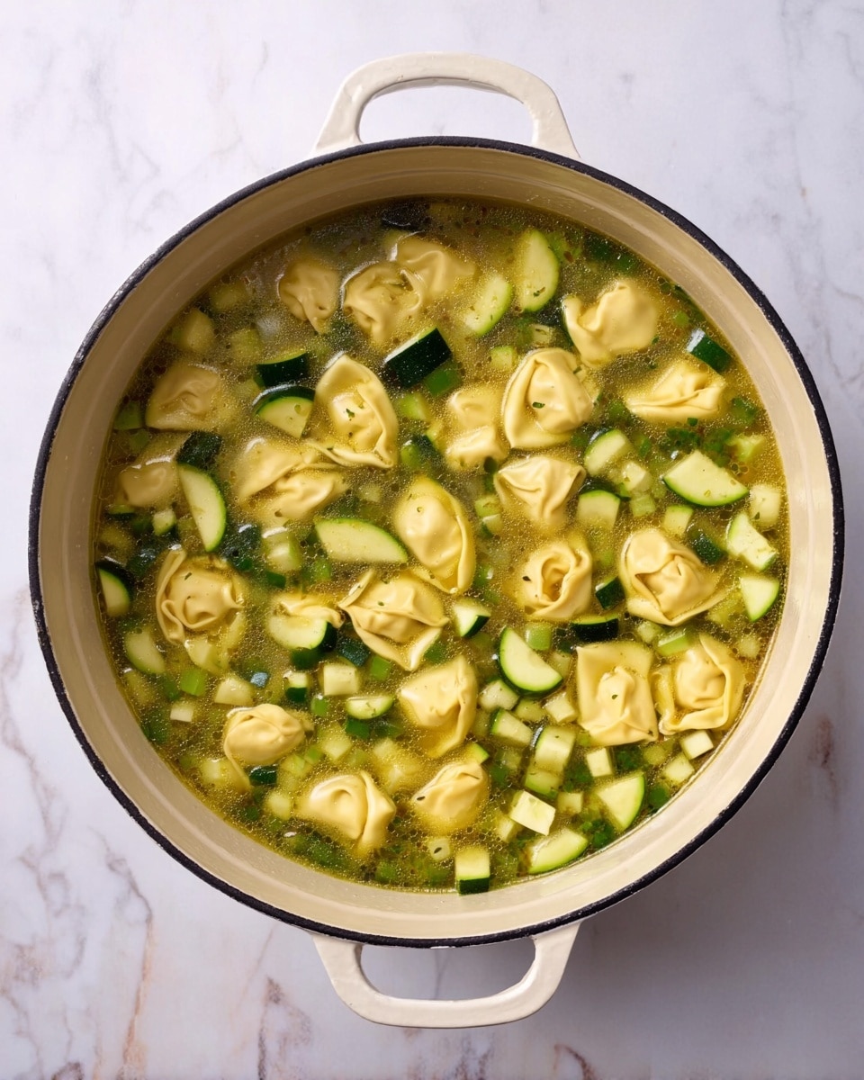 A top view of a white pot filled with clear broth soup containing three main layers: the base layer is the light greenish broth, the middle layer consists of small diced green zucchini pieces scattered throughout, and the top layer includes yellowish tortellini pasta pieces floating evenly across the surface. The pot is placed on a white marbled surface photo taken with an iphone --ar 4:5 --v 7