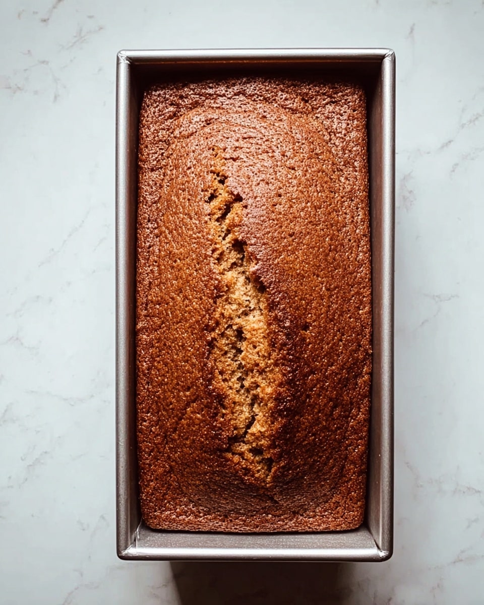 A single rectangular loaf of brown cake with a lightly cracked top surface sits inside a silver metal loaf pan. The cake has a slightly rough texture and a warm, baked color with darker brown edges all around. The background is a white marbled texture. photo taken with an iphone --ar 4:5 --v 7