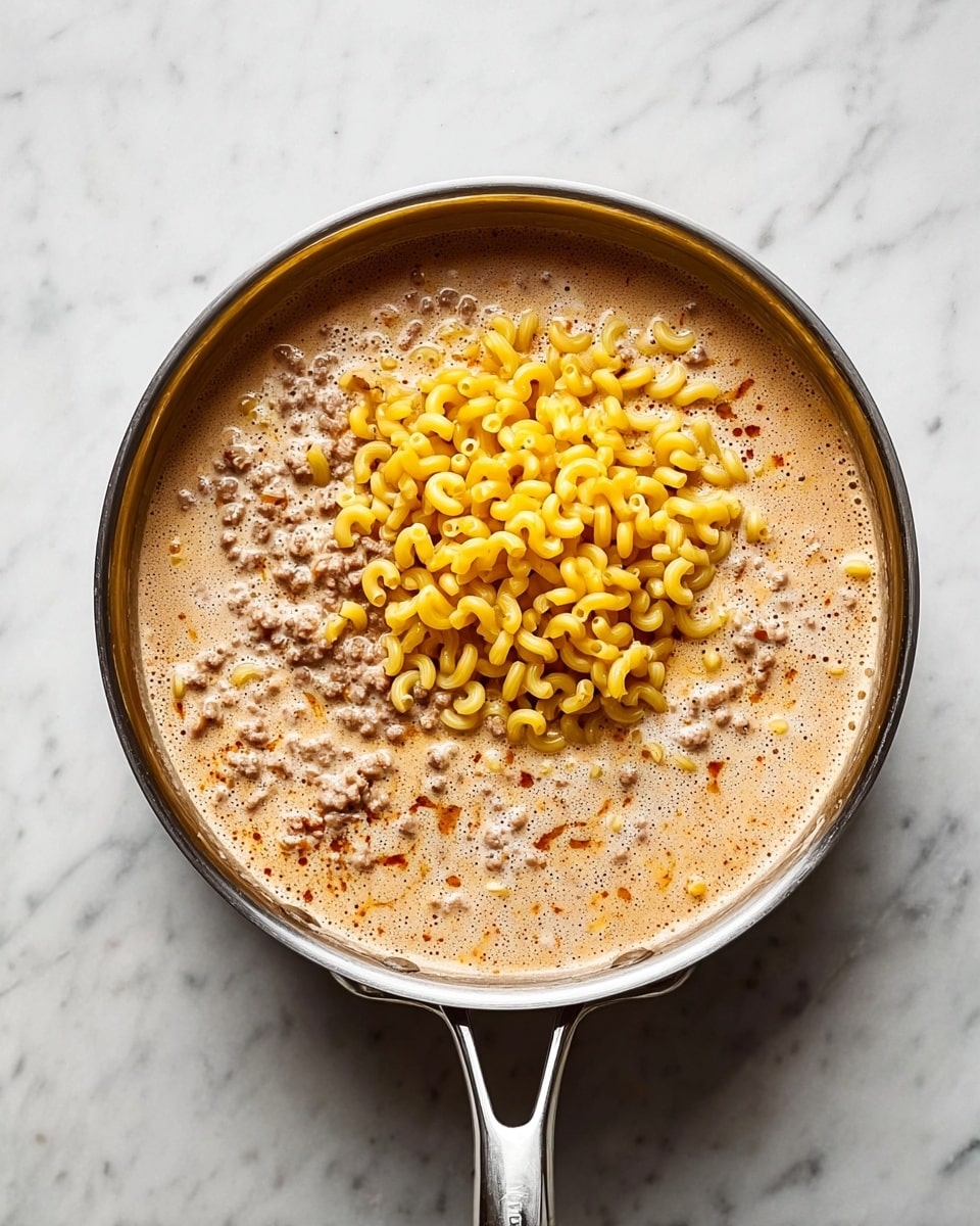 A shiny silver pot with a flat handle on a white marbled surface holds a creamy light brown soup mixed with small bits of ground meat, filling most of the pot. On top, a mound of small, curved elbow macaroni in a bright yellow color rests in the middle, creating contrast with the soup's smooth texture. The scene is lit softly from above, highlighting the bubbling foam and slight sheen on the soup’s surface. photo taken with an iphone --ar 4:5 --v 7