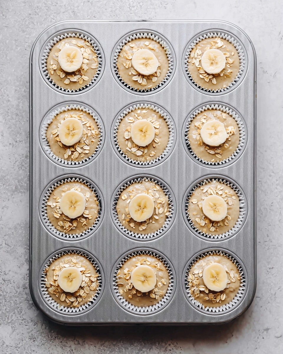 A metal muffin tray holds twelve white cupcake liners filled with light beige muffin batter. Each liner has one round, thin slice of banana placed on top, centered, with a small sprinkle of pale, raw oats arranged next to the banana slice. The tray is set on a white marbled textured surface. photo taken with an iphone --ar 4:5 --v 7