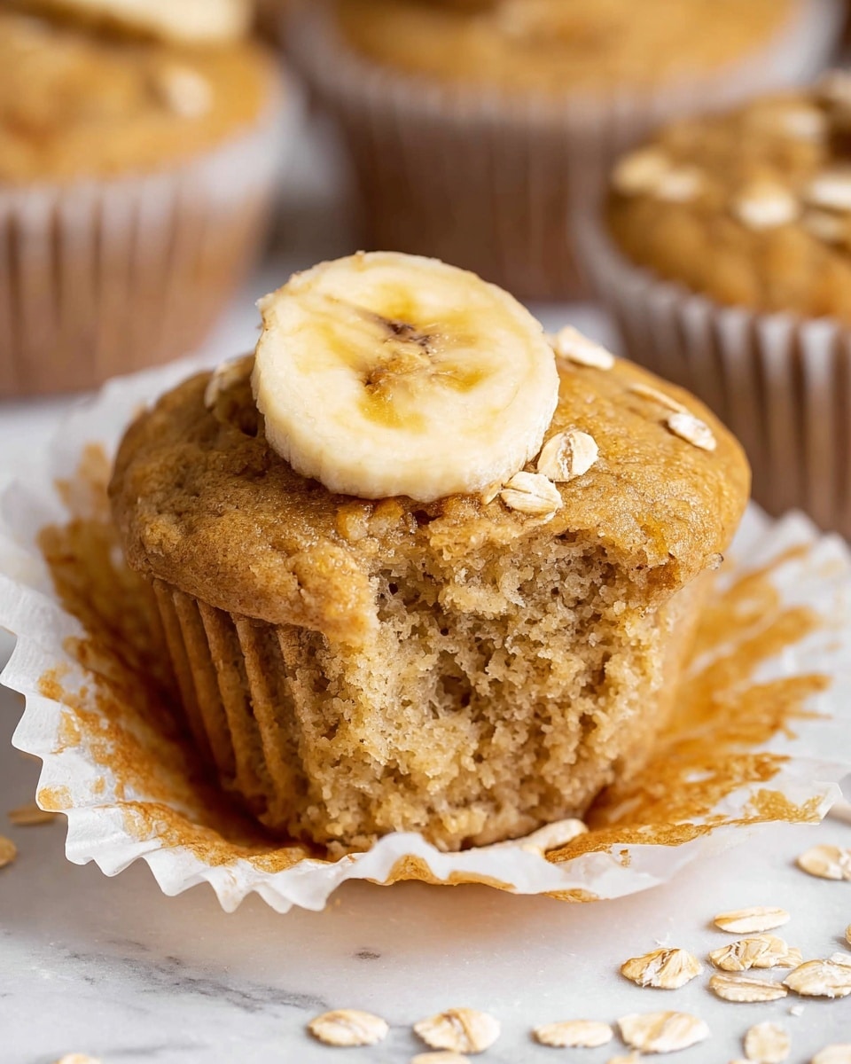 A single light brown muffin with a soft, moist texture is shown with its white paper liner partially peeled away, revealing the crumbly inside. On top, there is one round slice of banana with dark brown spots, and a small cluster of light beige rolled oats scattered nearby. The muffin sits on a white marbled surface with other muffins slightly blurred in the background. Photo taken with an iphone --ar 4:5 --v 7