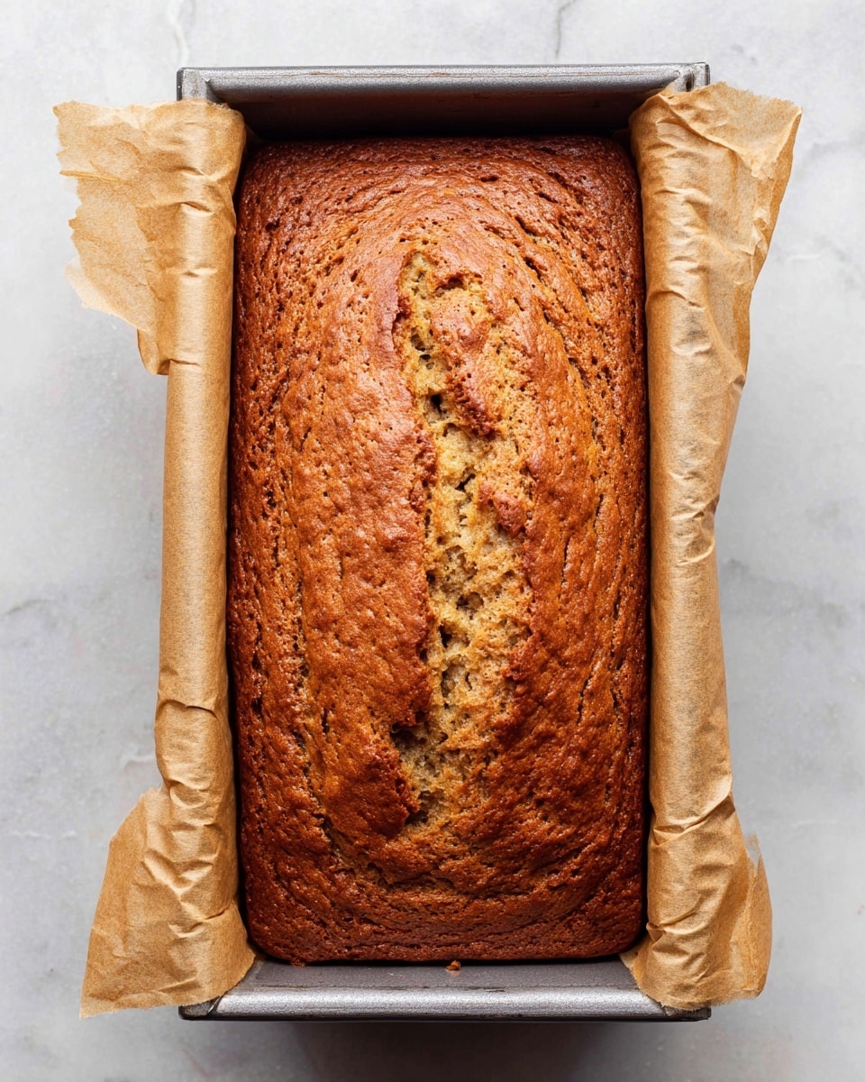 A golden brown loaf cake sits inside a metal baking pan lined with beige parchment paper that extends over the sides. The top of the cake is gently cracked with a textured surface showing small air pockets and a slightly shiny finish. The pan rests on a white marbled surface, enhancing the warm, rich color of the cake. Photo taken with an iphone --ar 4:5 --v 7
