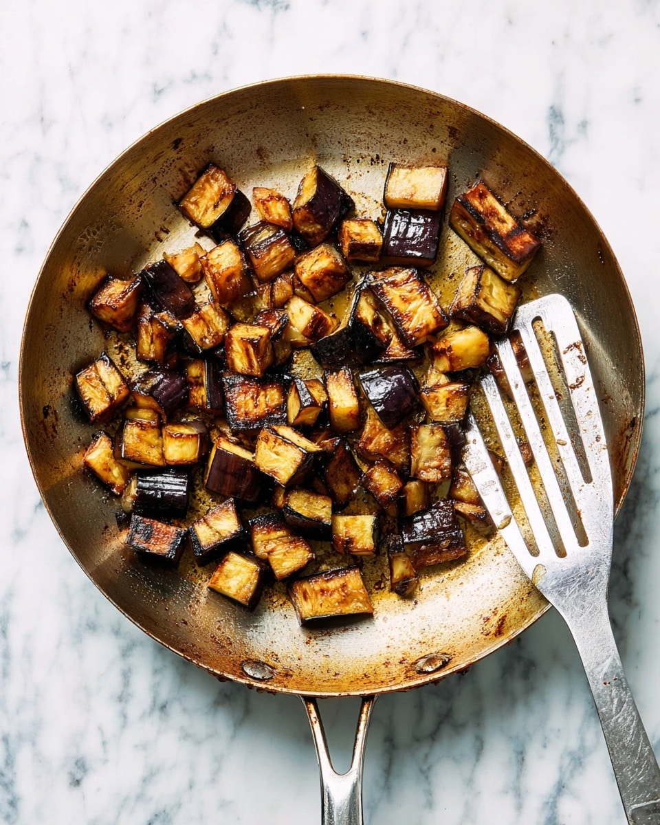 The image shows a white pan with a metal handle placed on a white marbled surface. Inside the pan are several pieces of cooked eggplant, cut into uneven cubes of different sizes. The eggplant pieces have a dark brown to black char on several sides, showing a mix of golden yellow insides and dark purple skin. A metal spatula with slots is resting in the pan on the right side, partly under the eggplant pieces. The pan’s surface has browned bits and food marks from cooking the eggplant, showing a slightly greasy texture. Photo taken with an iphone --ar 4:5 --v 7