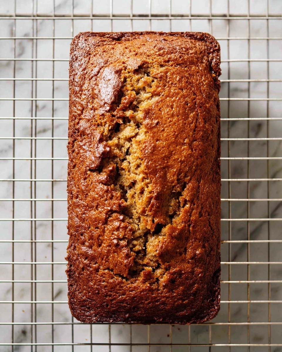 A loaf of banana bread is placed on a cooling rack above a white marbled surface. The bread has one visible layer with a cracked top crust showing a moist, grainy interior beneath. The crust is a rich golden brown with darker brown spots, and the texture looks dense yet soft. The cooling rack's silver grid contrasts with the warm tones of the bread, highlighting its shape and texture. photo taken with an iphone --ar 4:5 --v 7