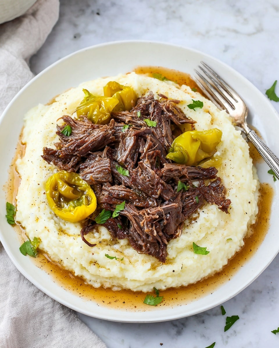 A white plate holds a layer of creamy mashed potatoes, white with a smooth texture and slight lumps, spread evenly in a circular shape. On top is a generous pile of shredded dark brown beef, moist and tender with some pieces showing a fibrous texture. Bright yellow-green whole peppers are placed on the mashed potatoes around the beef, adding contrast and color. Small green parsley leaves are sprinkled lightly over the meat and mashed potatoes. A light brown sauce with a thin, glossy texture pools slightly around the edges of the mashed potatoes. The plate sits on a white marbled surface, with a silver fork resting on the right side of the plate. Photo taken with an iphone --ar 4:5 --v 7