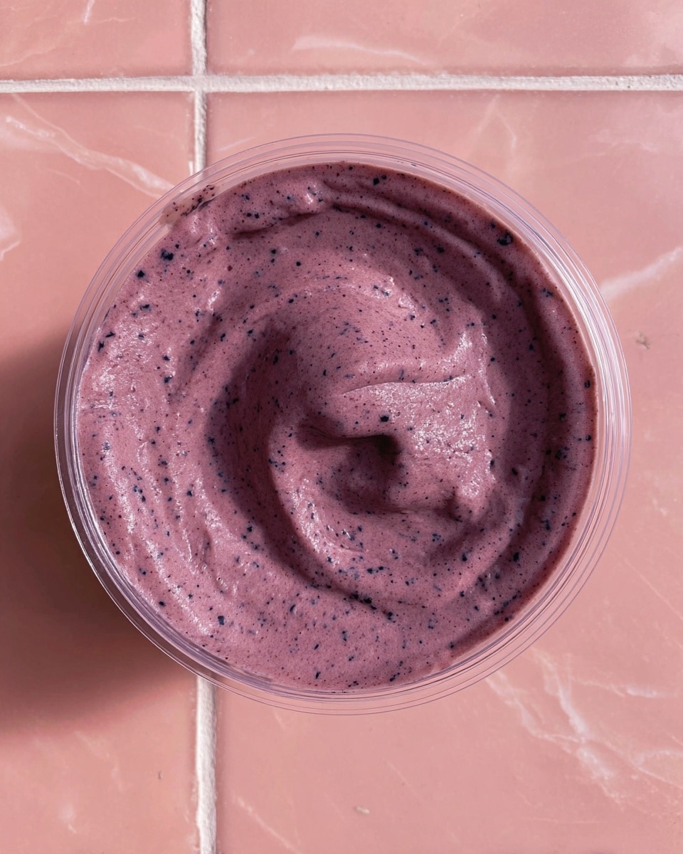 A single layer of thick, creamy purple smoothie fills a clear round container, showing a smooth yet slightly textured surface with tiny darker purple specks mixed in. The container's lid edges are visible at the top, and it sits on a pink tiled surface. The creamy mixture is swirled gently in the center, creating soft folds and light shadows that add depth. The background is replaced with a white marbled texture. photo taken with an iphone --ar 4:5 --v 7