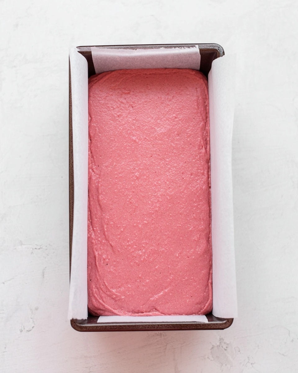 A rectangular metal baking pan lined with white parchment paper holds a smooth, thick layer of bright pink batter evenly spread inside. The batter has a soft, velvety texture with tiny air bubbles visible on the surface, filling the pan completely from edge to edge. The pan is placed on a white marbled textured surface. photo taken with an iphone --ar 4:5 --v 7
