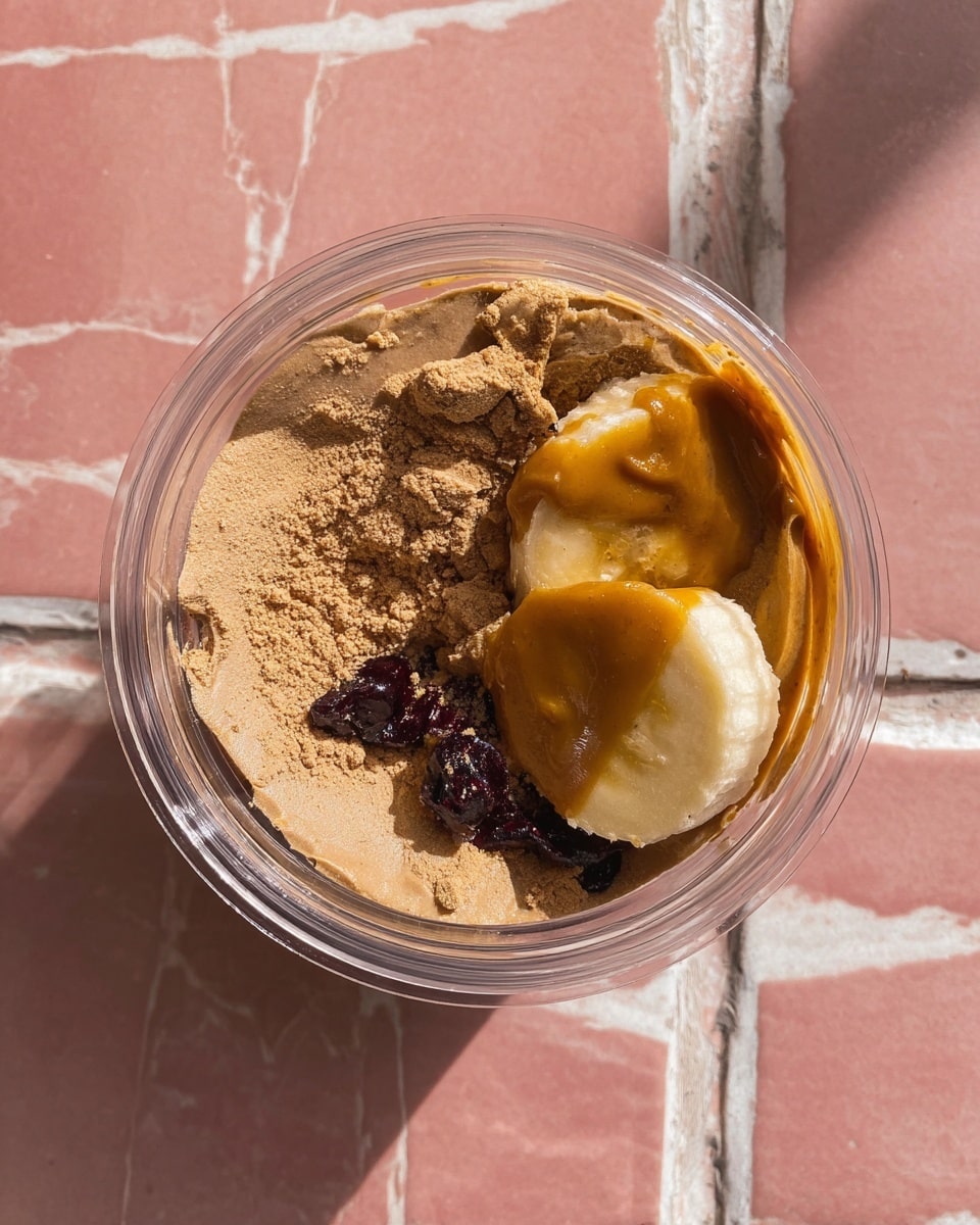 A clear round container filled with three main layers: a light brown powder covering about half the space, a light yellow banana slice partly covered with a thick, smooth, caramel-colored spread, and a few small dark purple berries mostly hidden under the banana and spread. The container sits on a white marbled surface with worn pink tiles in the background. The light highlights the textures clearly, showing the powder's grainy look, the banana's softness, and the spread's shiny creaminess. photo taken with an iphone --ar 4:5 --v 7