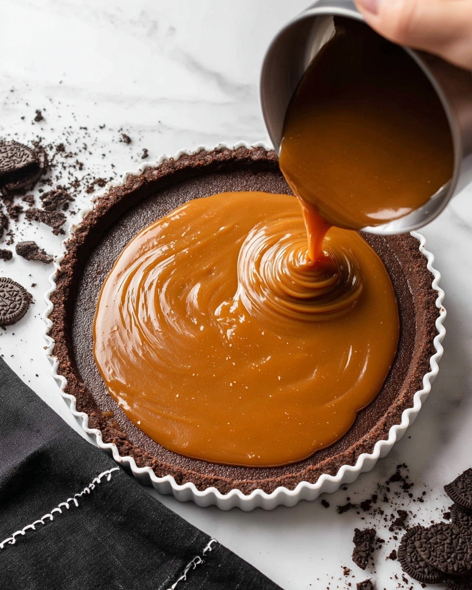 A chocolate crust tart shell in a white tart pan sits on a white marbled surface with some broken chocolate cookie crumbs around it. A woman's hand pours a thick, smooth, and glossy caramel layer from a silver pot onto the inside of the crust, creating shiny swirls as it fills the center. The rich brown crust has a crumbly texture with tall edges holding the warm caramel. Nearby, a woman's hand holds the pot steady as the caramel flows into the tart shell. Photo taken with an iphone --ar 4:5 --v 7