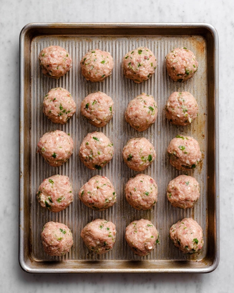 The image shows a metal baking tray filled with sixteen raw meatballs arranged in rows. Each meatball is light pink in color with small green herb pieces and tiny white and red bits mixed throughout. The meatballs have a smooth but slightly textured surface and are evenly spaced on the tray, which has a ridged pattern and slight discoloration from use. The background is a white marbled texture. photo taken with an iphone --ar 4:5 --v 7