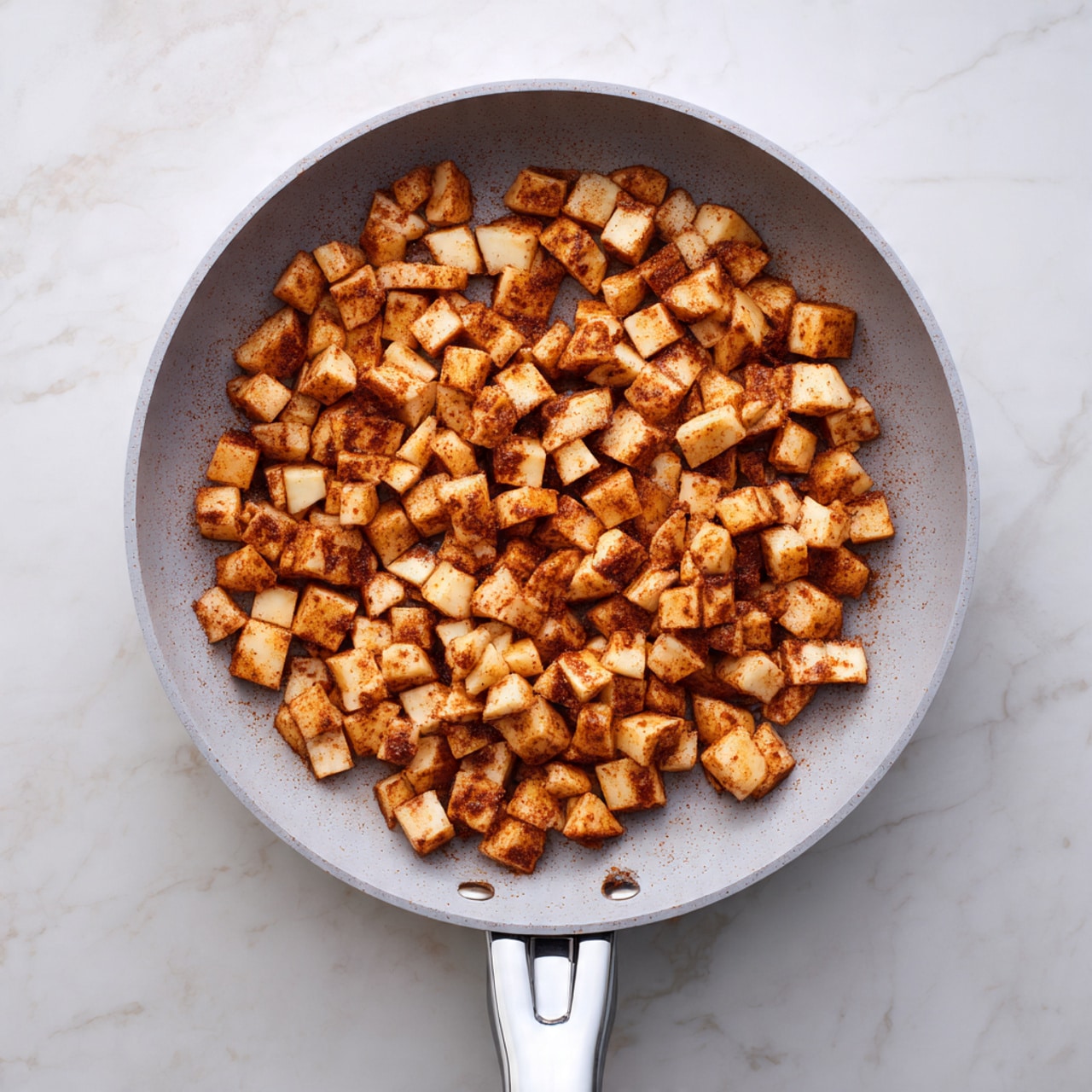 A gray pan with two handles sits on a white marbled surface, filled with small, evenly cut, golden-brown cubes of cooked food, lightly coated with a reddish-brown spice. The cubes are spread in a single layer, showing a slightly crispy texture. The pan’s light gray handle extends toward the bottom right corner of the image. photo taken with an iphone --ar 4:5 --v 7