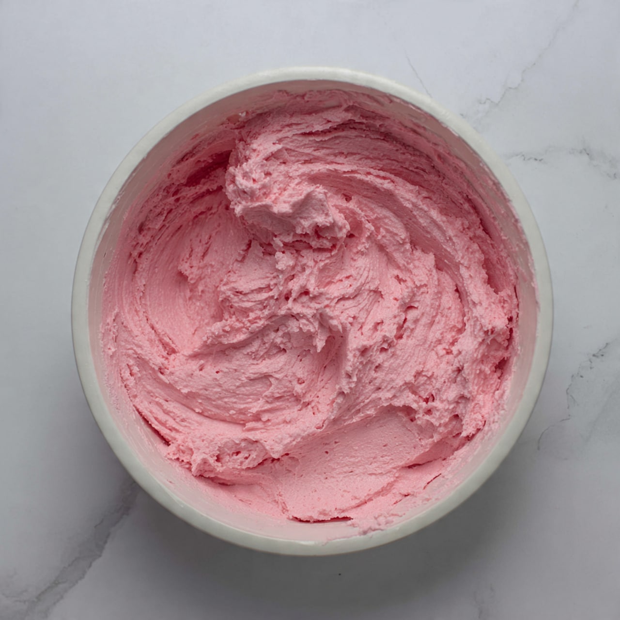 A white bowl filled with a thick, fluffy pink mixture that has a soft texture with swirls and peaks. The mixture is evenly spread inside the bowl, with some of it sticking to the sides. The bowl sits on a white marbled surface. photo taken with an iphone --ar 4:5 --v 7