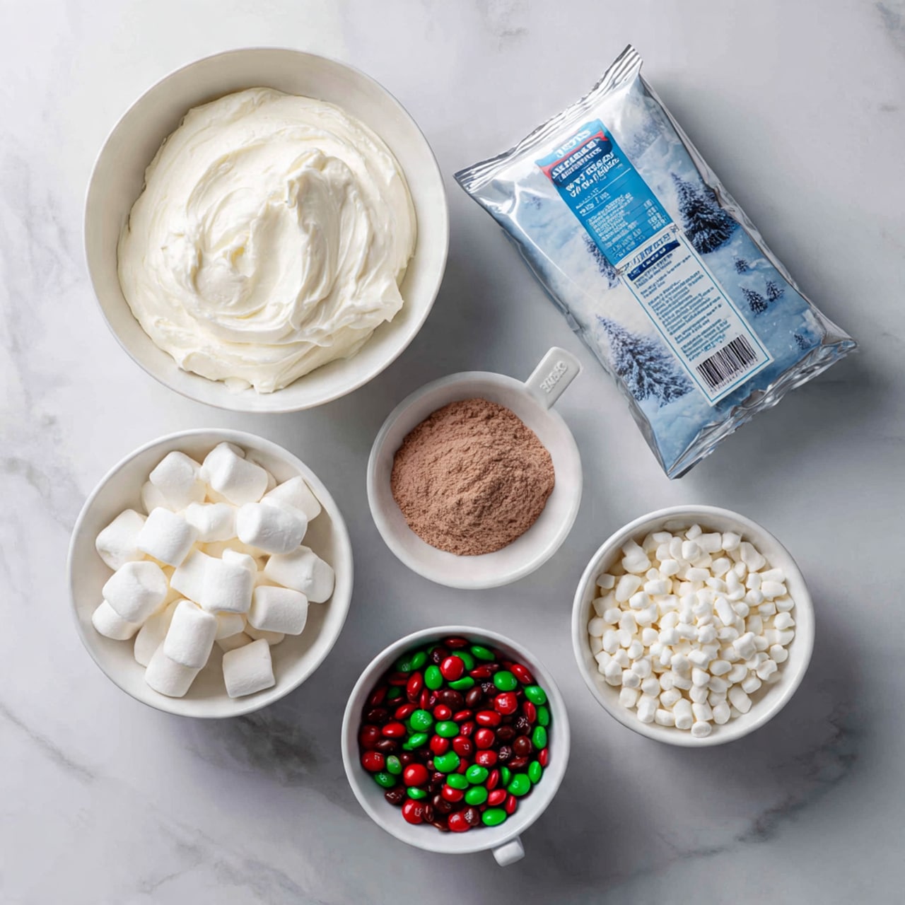 A top-down view of seven baking ingredients arranged on a white marbled surface: a white bowl filled with smooth white cream, a white measuring cup filled with fine light brown powder, a small white bowl with mini white marshmallows, another small white bowl filled with tiny white beads (possibly mini marshmallows or puffed rice), and a white bowl with red and green candy-coated chocolates. A silver wrapped block of cream cheese is placed near the top right, and next to it is a white jar with a blue lid labeled