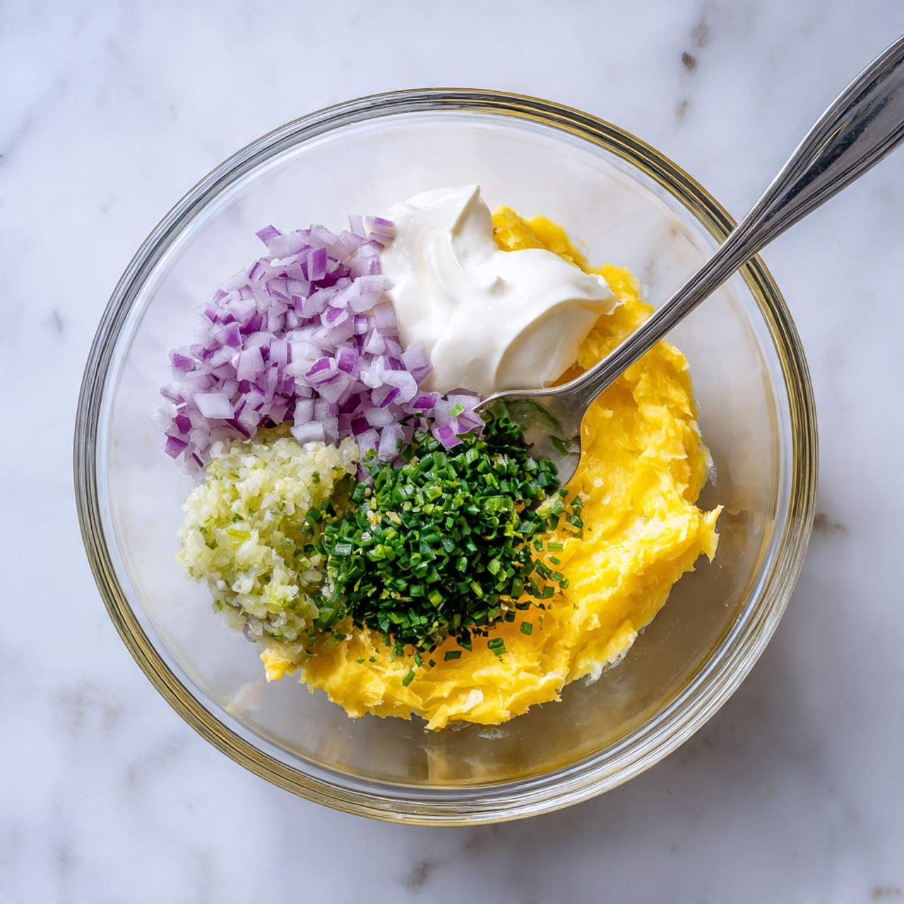 Smoked Salmon Deviled Eggs Recipe 5 A clear glass bowl sits on a white marbled surface, filled with several colorful ingredients layered loosely. At the bottom, there is a layer of bright yellow mashed yolks. On one side, there is a small pile of finely chopped light purple onions. Next to it is a small grouping of green herbs, finely chopped. There is a dollop of thick white mayonnaise placed on top of the yellow yolks. A small portion of pale green relish is positioned near the onions. A stainless steel fork with a silver handle rests inside the bowl, leaning against the side. Photo taken with an iphone --ar 4:5 --v 7