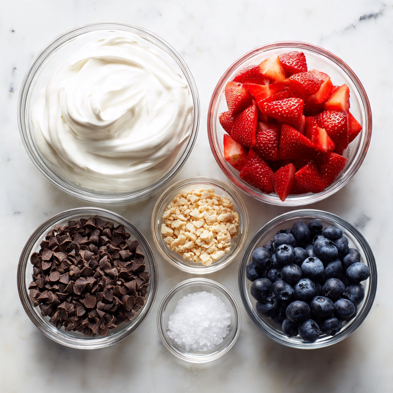 A white plate sits on a red mat with a white marbled background. On the plate are nine clusters made of dark brown chocolate with a rough texture. Two clusters are broken in half at the front, showing a creamy white and pink filling inside with visible whole blueberries and bits of red fruit. Scattered blueberries are between the clusters. Three fresh strawberries with green leaves are placed around the clusters near the edges of the plate. A few flakes of salt or sugar crystals are sprinkled on top of the chocolate clusters. photo taken with an iphone --ar 4:5 --v 7