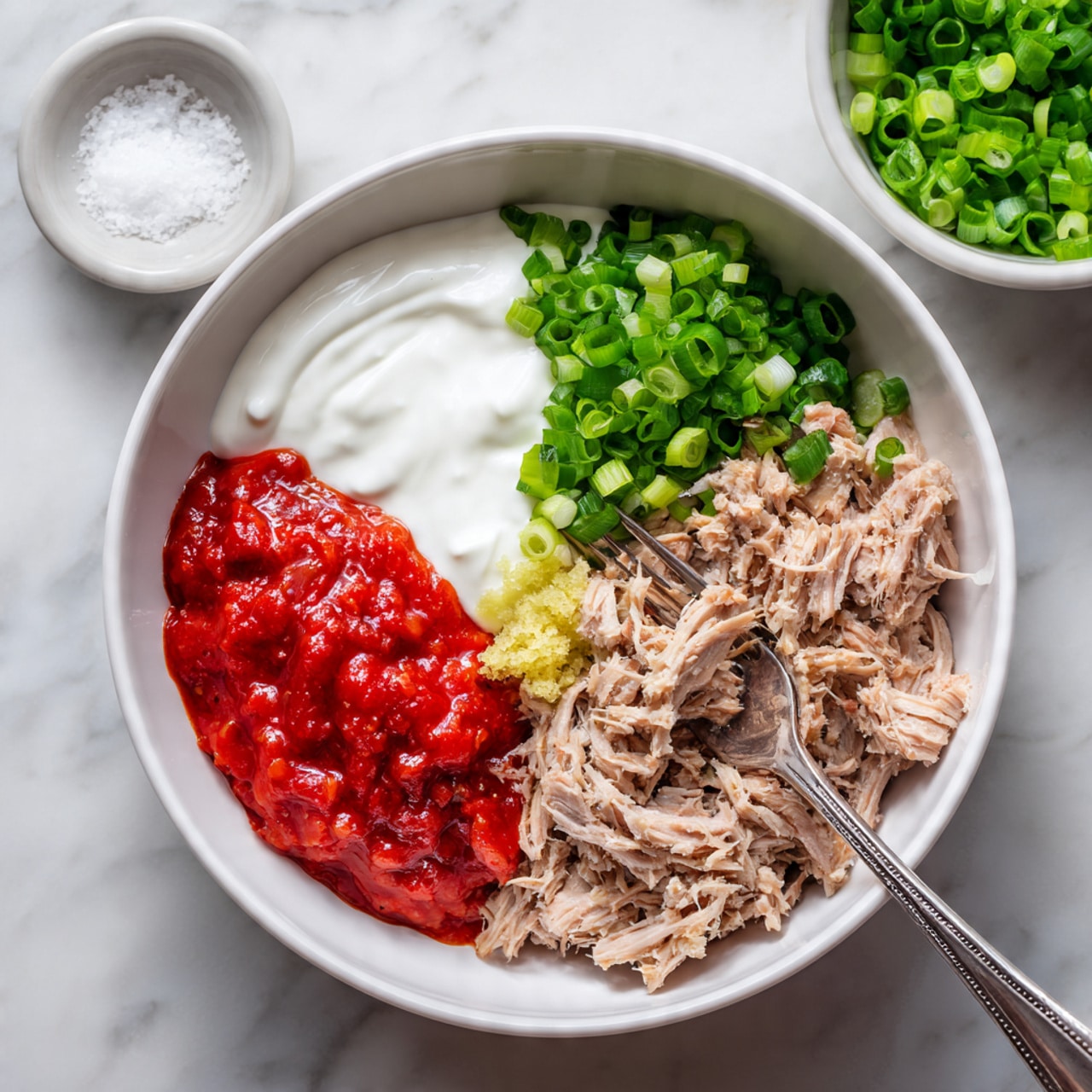 In the white bowl, there are three main layers arranged side by side: on the left, a smooth white yogurt layer; next to it, a bright red sauce with a thick texture; and on the right, light brown chunks of shredded tuna mixed with a small amount of grated yellow ginger near the front. A silver fork rests inside the bowl, touching the tuna. The bowl sits on a white marbled surface, and in the top left corner, there is a small round white bowl of coarse salt, while at the bottom right edge, there is a partial view of a white bowl filled with sliced green onions. Photo taken with an iphone --ar 4:5 --v 7