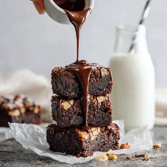A stack of three dark brown brownies with a rich texture, each showing chunks of lighter nuts inside, sits on white parchment paper over a rustic patterned metal surface. A thin stream of shiny dark chocolate sauce is being poured from above by a woman's hand, covering the top and dripping down the sides of the brownies. Behind the stack, there is a glass bottle filled with white milk, all set on a white marbled surface with a soft, bright background. Photo taken with an iphone --ar 4:5 --v 7