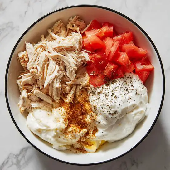 A white bowl with a black rim sits on a white marbled surface. Inside the bowl, five main layers are arranged without mixing: shredded light brown cooked chicken at the top left, a smooth white creamy layer of sour cream with black pepper on the top right, small chopped bright red tomatoes on the bottom left, pale yellow mayonnaise next to the sour cream on the bottom right, and a pile of light brown spice powder between the tomatoes and mayonnaise. The layers are neatly placed in separate patches but close together, showing distinct textures and colors. Photo taken with an iphone --ar 4:5 --v 7
