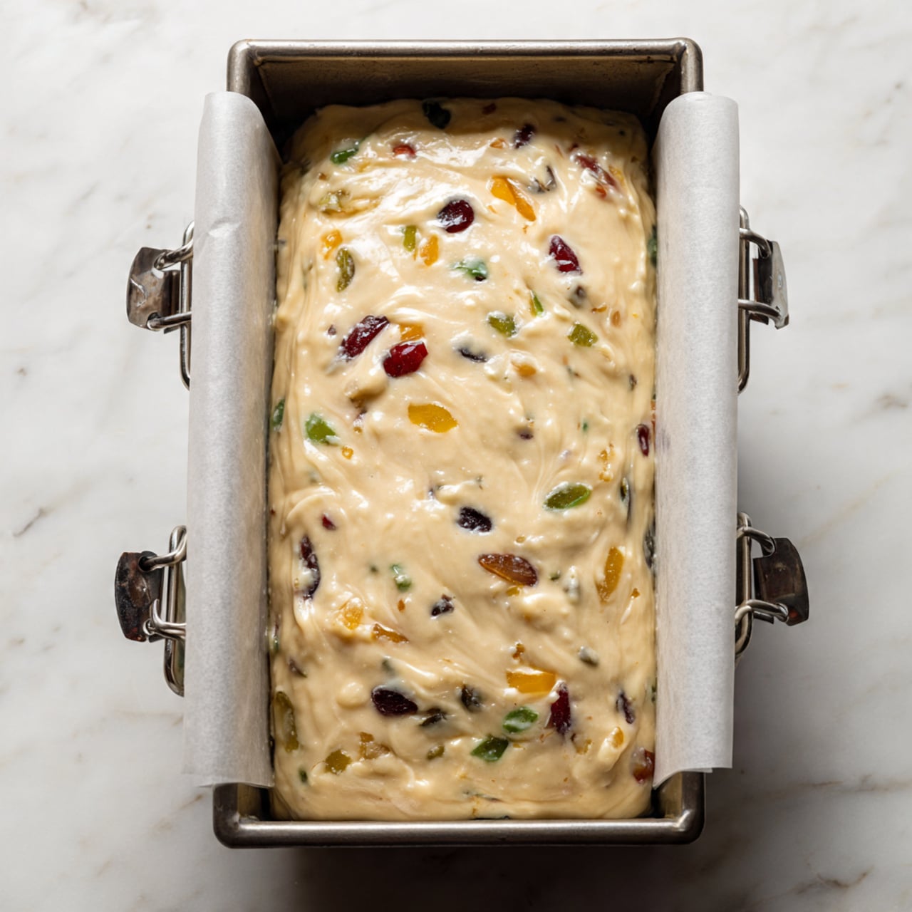 The image shows a metal loaf pan lined with white parchment paper held in place by two metal clips on either side. Inside the pan is a smooth, thick batter that is pale cream in color, speckled with pieces of yellow, green, red, and dark purple fruits or nuts spread evenly throughout. The batter fills almost to the top of the pan, with a slightly uneven surface and some gentle swirls. The pan sits on a white marbled surface. photo taken with an iphone --ar 4:5 --v 7