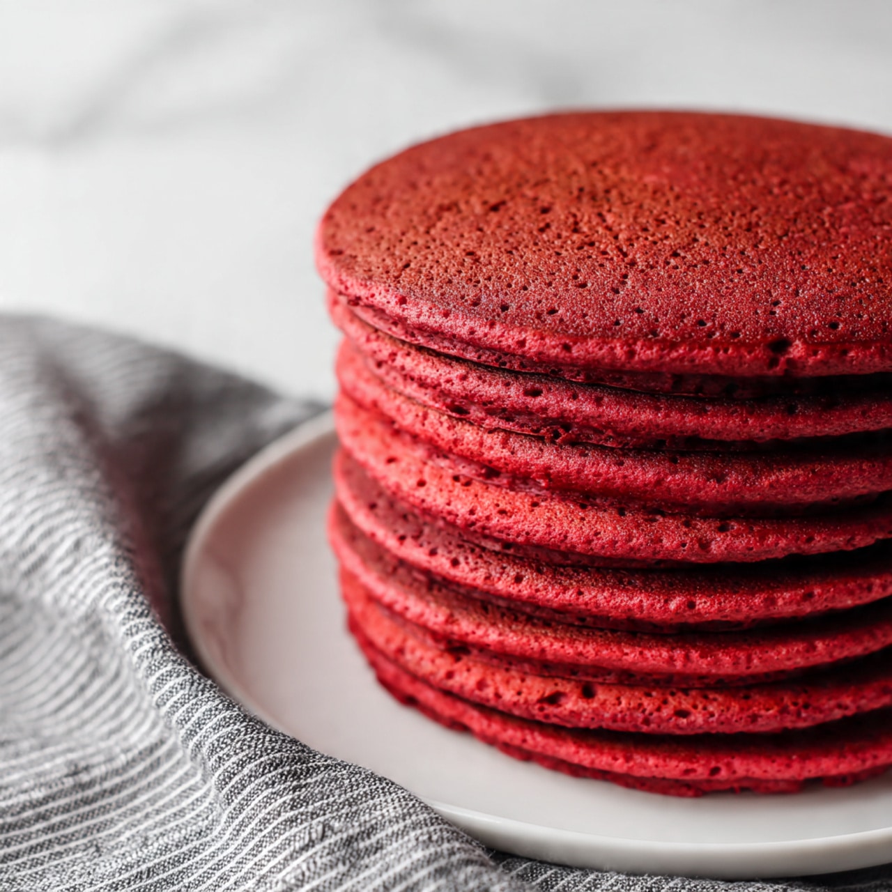 A tall stack of seven thick, round red pancakes sits on a white plate, each pancake showing a spongy texture with small air holes on the sides, giving them a slightly rough surface. The pancakes are deep ruby red with a moist look, and the stack is centered in the image, placed on a white marbled surface. The edges of the pancakes are slightly uneven, showing their softness and freshness, with no toppings or syrups on them. Photo taken with an iphone --ar 4:5 --v 7