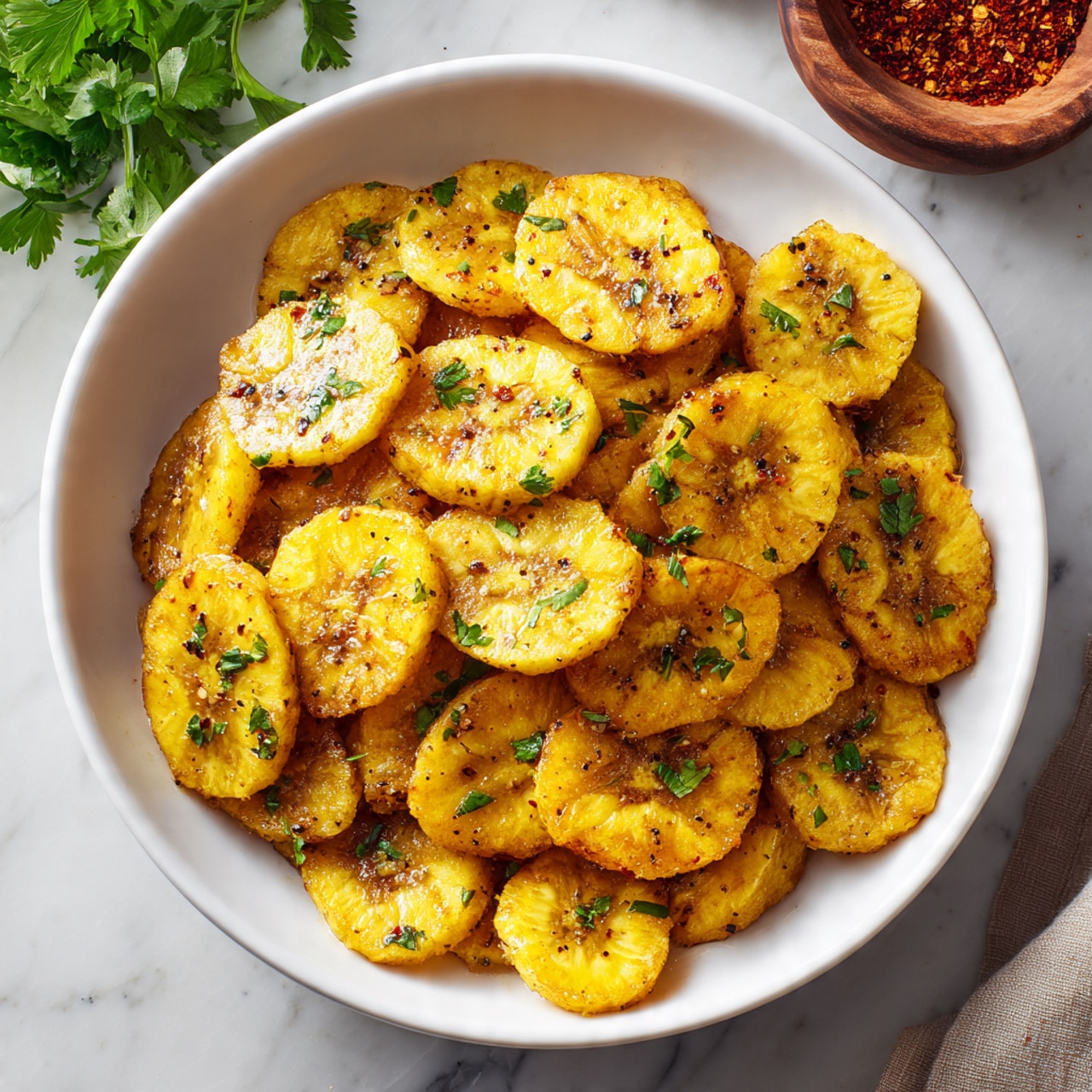 A close-up view of a small brown bowl filled with chunky bright green guacamole that has a slightly textured surface with visible bits of herbs mixed in, positioned in the front center. A woman's hand with blue nail polish is dipping a single round, golden-brown plantain chip with a slightly crispy and uneven surface into the guacamole. Behind, there is a larger bowl filled with more plantain chips that have deep golden to dark brown hues and a rough texture. The setting is on a white marbled surface with fresh green parsley and red chili flakes in small wooden bowls scattered around, along with a wooden kitchen utensil and a white cloth with red stitching detail. Photo taken with an iphone --ar 4:5 --v 7