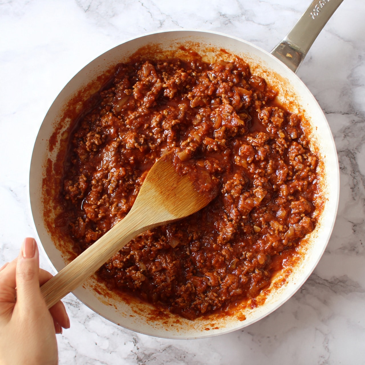 The image shows a white baking dish on a white marbled surface with a layered dish inside. The bottom layer is a bed of shredded orange cheddar cheese spread unevenly across the base, with some pieces scattered around the edges of the dish. On top of the cheese, there is a layer of cooked ground meat mixed with a reddish-brown sauce, visible mostly around the edges. The top layer consists of twelve soft, golden-brown slider buns arranged closely in a 3x4 grid, with the shiny tops showing natural wrinkles and slight cracks. The buns sit neatly together, covering most of the meat and cheese inside. photo taken with an iphone --ar 4:5 --v 7