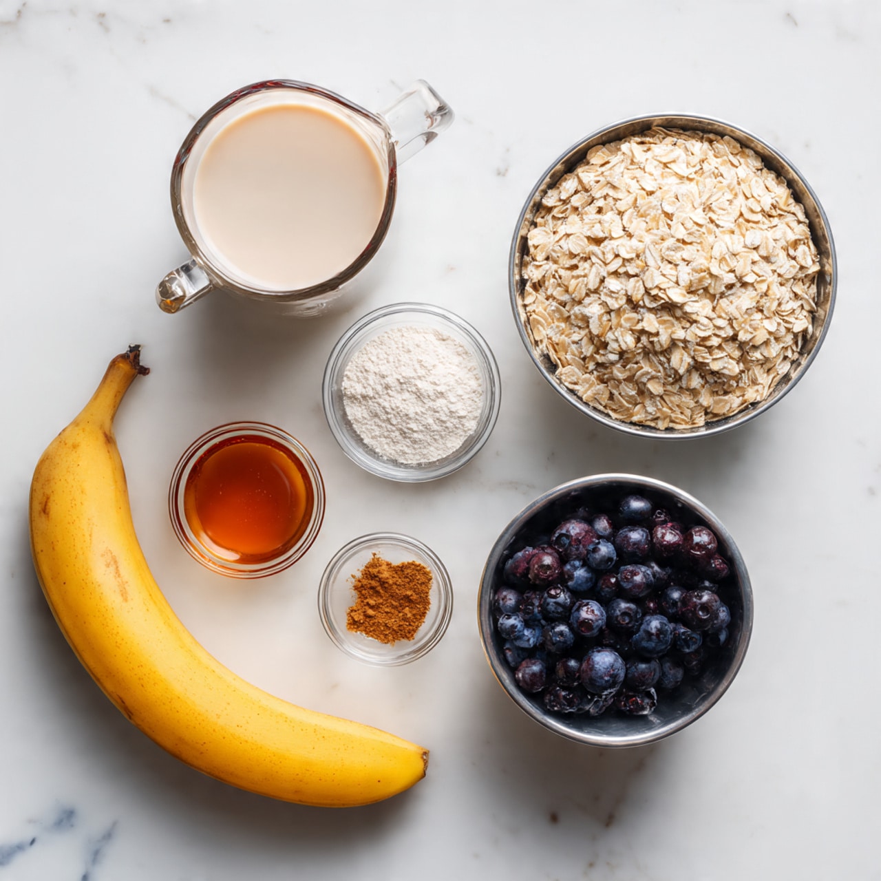 The image shows six ingredients placed neatly on a white marbled surface: a clear measuring cup filled with light beige almond milk on the left, a metal cup filled with beige rolled oats on the upper right, a small round glass bowl with white baking powder, a small glass bowl with light brown powder (likely a spice), a small glass bowl with amber-colored maple syrup, a ripe yellow banana with a bit of brown spots near the center, and a metal cup filled with dark purple frozen blueberries on the lower right, all arranged with clear space between them photo taken with an iphone --ar 4:5 --v 7