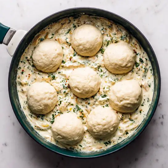 A dark green skillet filled with a creamy white sauce mixed with small bits of chicken and herbs, giving the sauce a speckled look. On top, there are eight round biscuits with soft, pale golden tops, evenly spaced, sitting in the sauce. The skillet is placed on a white marbled surface, and the sauce fills the pan almost to the edge. photo taken with an iphone --ar 4:5 --v 7