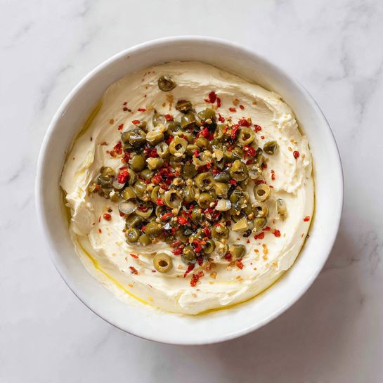 Inside a white bowl on a white marbled surface, there are six different ingredients neatly placed in separate sections. In the top left, there are white cream cheese cubes with a soft texture. To the right, there is a smooth mound of white sour cream. Below the cream cheese, there is a pile of finely shredded pale yellow cheese. Near the center, there is a small mound of beige powder. Next to it, some coarse white salt is placed. Finally, there is a sprinkling of black pepper near the salt. The ingredients are arranged carefully without mixing. Photo taken with an iphone --ar 4:5 --v 7