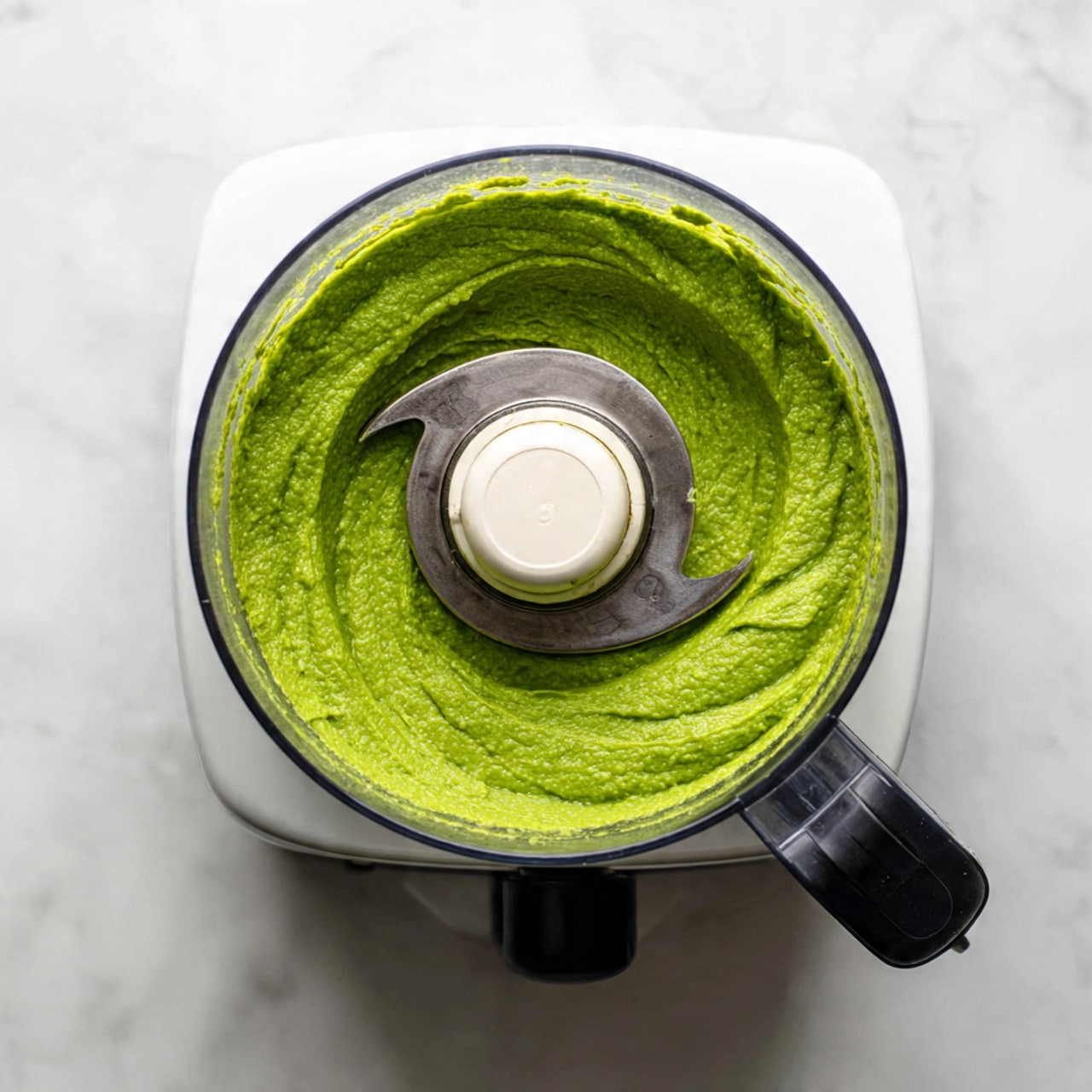 The image shows a white food processor sitting on a white marbled surface. Inside the clear bowl of the food processor, there is a smooth, bright green paste that covers the blade and the bottom of the bowl evenly. The blade is white and silver, mostly covered by the green paste. The food processor has a black and silver handle on the front. There are no extra layers in the image, just the single layer of green paste inside the clear bowl. Photo taken with an iphone --ar 4:5 --v 7