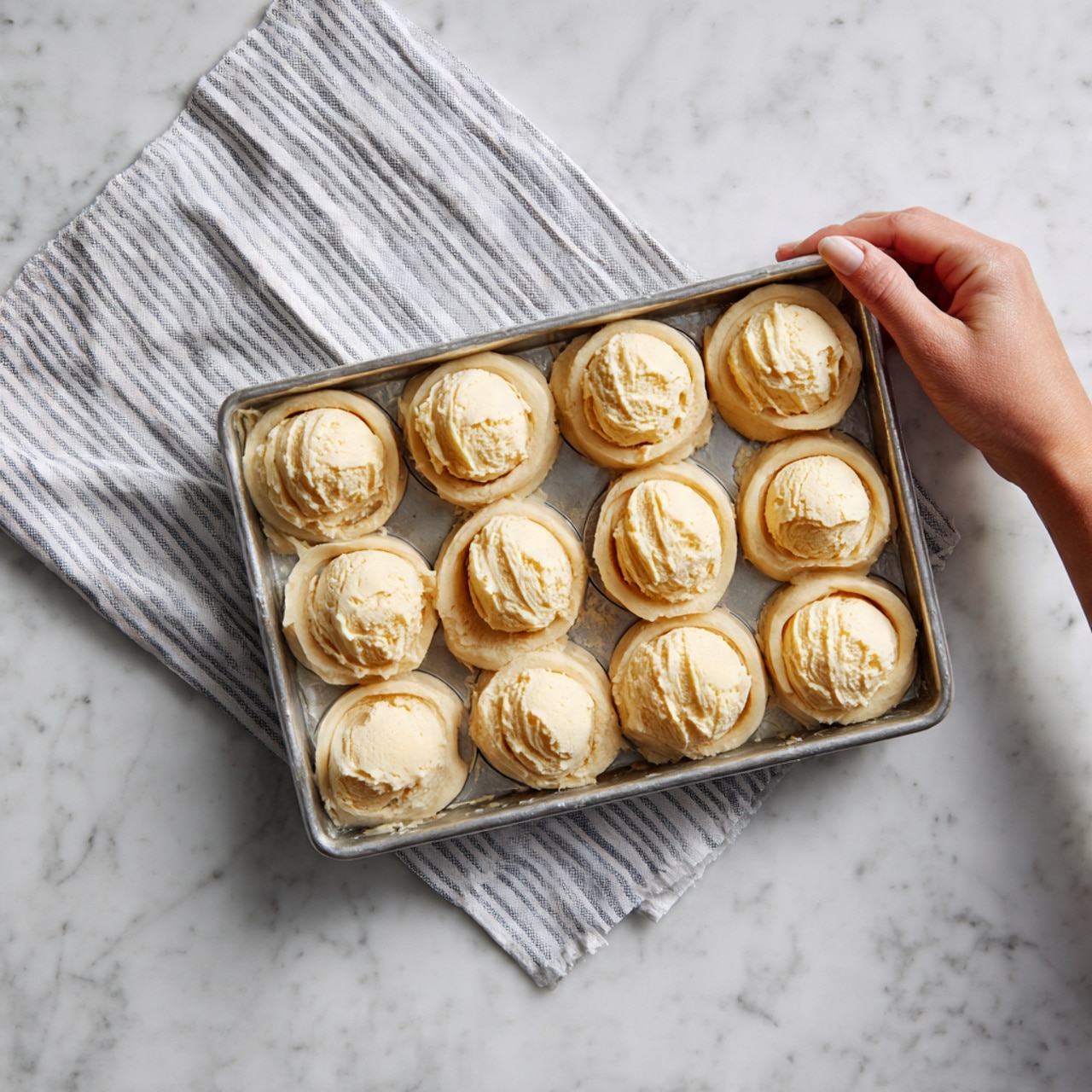 The image shows a muffin tray with twelve white paper linings shaped like flowers, each holding a round ball of dough inside. The dough is light beige and smooth, resting in the middle of each paper lining. The tray is silver and sits on a white marbled surface, giving the whole scene a clean and neat look. Photo taken with an iphone --ar 4:5 --v 7