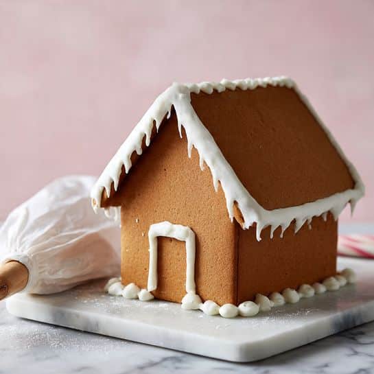 A small gingerbread house is placed on a white marble board with a pale pink background. The house has four visible flat brown gingerbread walls and two slanted brown gingerbread roof panels attached with thick white icing at the edges and corners. The icing is also piped along the base where the walls meet the board. To the left of the house, there is a filled white pastry bag lying on the board, ready for more icing work. The overall image shows the simple structure and raw look of the gingerbread house before decoration photo taken with an iphone --ar 4:5 --v 7