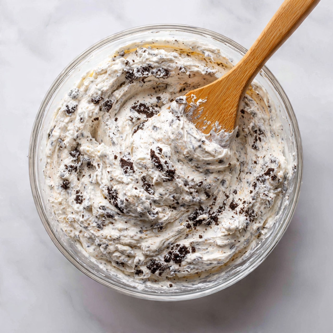 A clear glass bowl filled with a thick, whipped mixture that is light gray with small, dark chocolate cookie pieces scattered evenly throughout. A wooden spatula is partially buried in the mixture, showing the texture as smooth, creamy, and fluffy with visible cookie crumbs mixed in. The bowl is placed on a white marbled surface, giving a clean, bright look to the scene. photo taken with an iphone --ar 4:5 --v 7