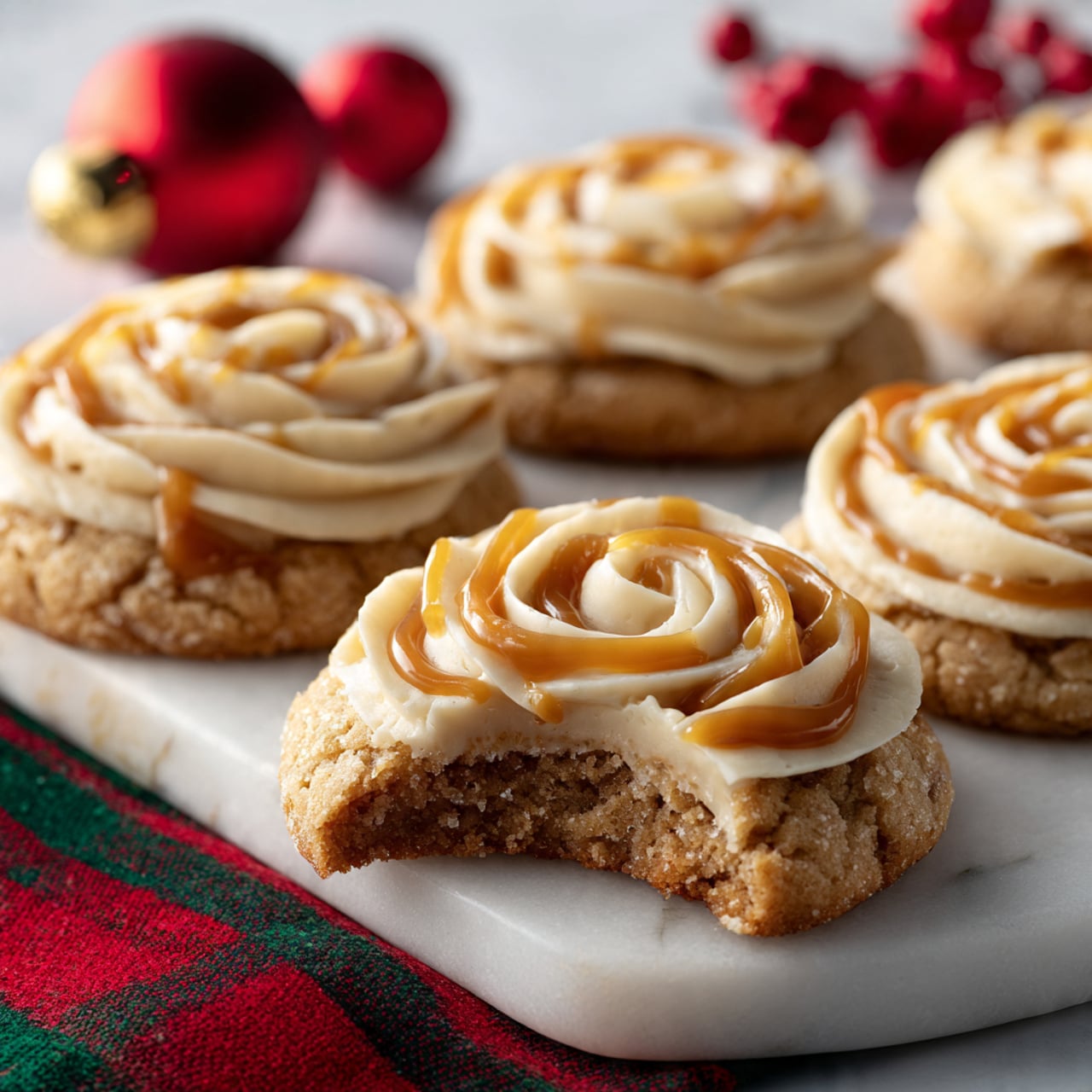 The image shows several soft brown cookies with slightly rough textures, each topped with two layers of creamy light beige frosting in swirled rose shapes. On top of the frosting, there is a smooth, golden caramel drizzle that adds a shiny highlight. One cookie is missing a bite, revealing its moist interior and the thick layer of frosting between the cookie halves. The cookies are placed on a white marbled surface, with parts of a red and green cloth and a small red Christmas ornament visible in the background. photo taken with an iphone --ar 4:5 --v 7