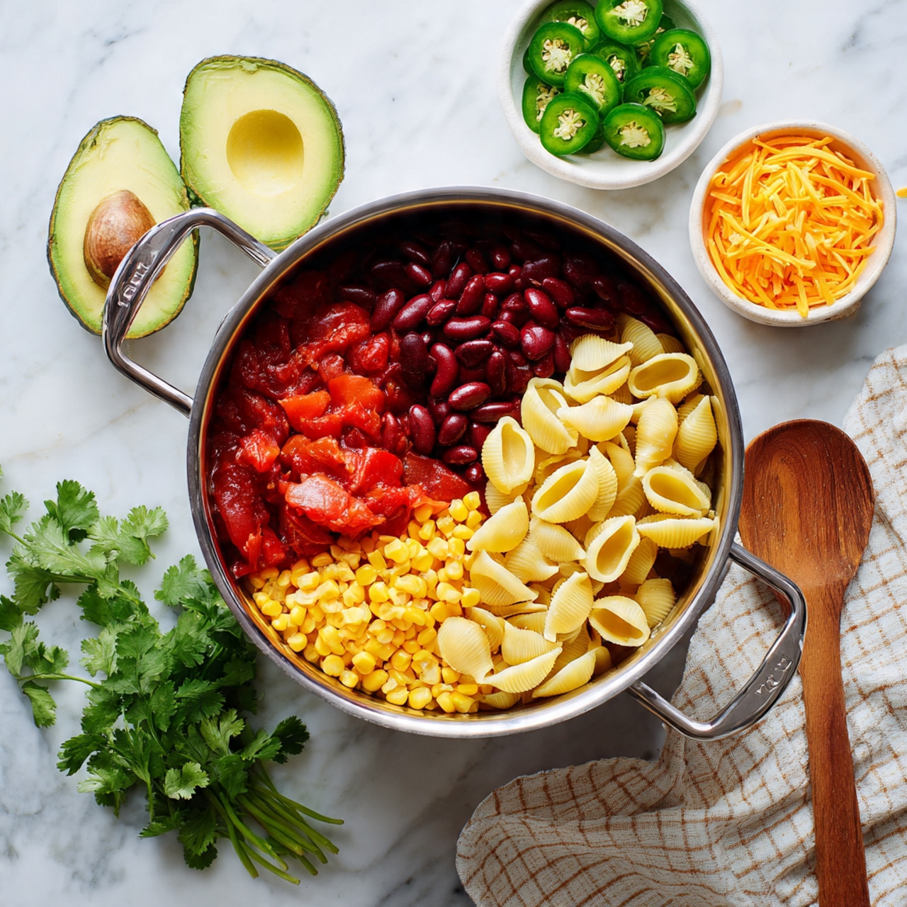 A round silver pan with a long handle holds a thick soup with visible chunks of red tomatoes, white beans, black beans, and small pasta pieces in a rich, reddish-orange broth. On top, there is a large dollop of white sour cream covering the center-left portion, and shredded yellow cheddar cheese scattered over the right side of the sour cream and soup. The pan is placed on a white marbled surface, surrounded by half an avocado with a brown seed, green chili peppers, a small white bowl filled with more shredded cheddar cheese, a wooden spoon, a beige and brown checkered cloth, and a small bunch of cilantro. photo taken with an iphone --ar 4:5 --v 7