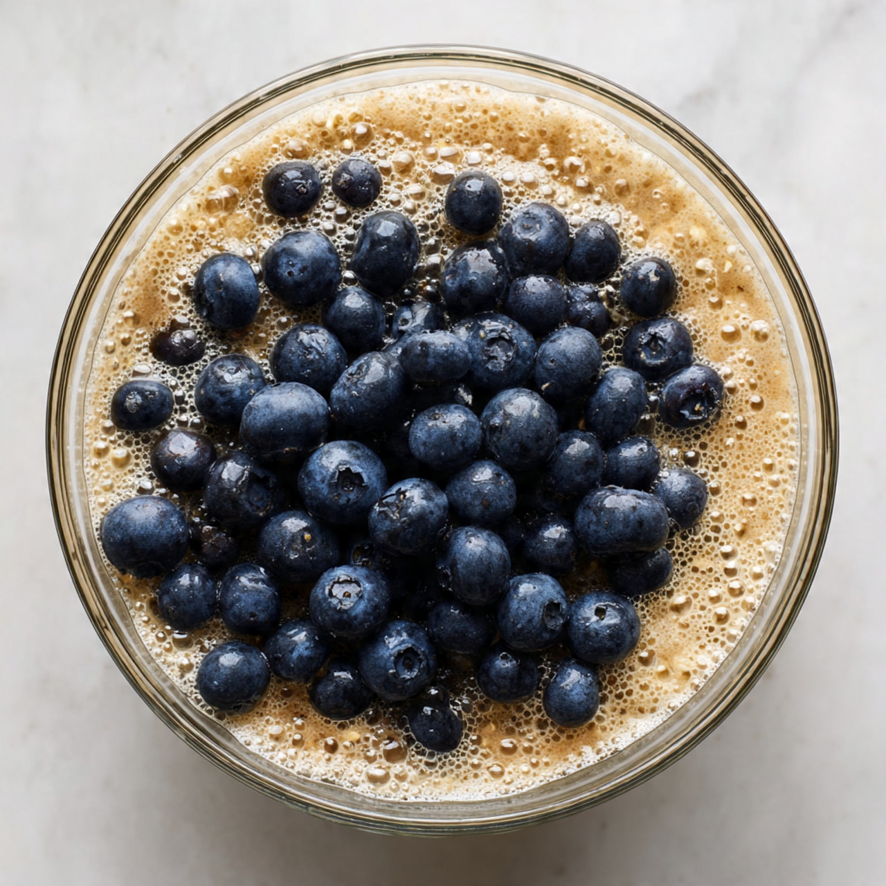 A clear glass bowl holds a mix of light brown soaked oats with visible bubbles on the surface, giving a slightly foamy texture. On top of the oats, there is a pile of fresh, plump blueberries that are deep blue to almost black in color, creating a strong contrast with the lighter oat layer underneath. The bowl is placed on a white marbled surface, enhancing the natural colors of the ingredients. photo taken with an iphone --ar 4:5 --v 7