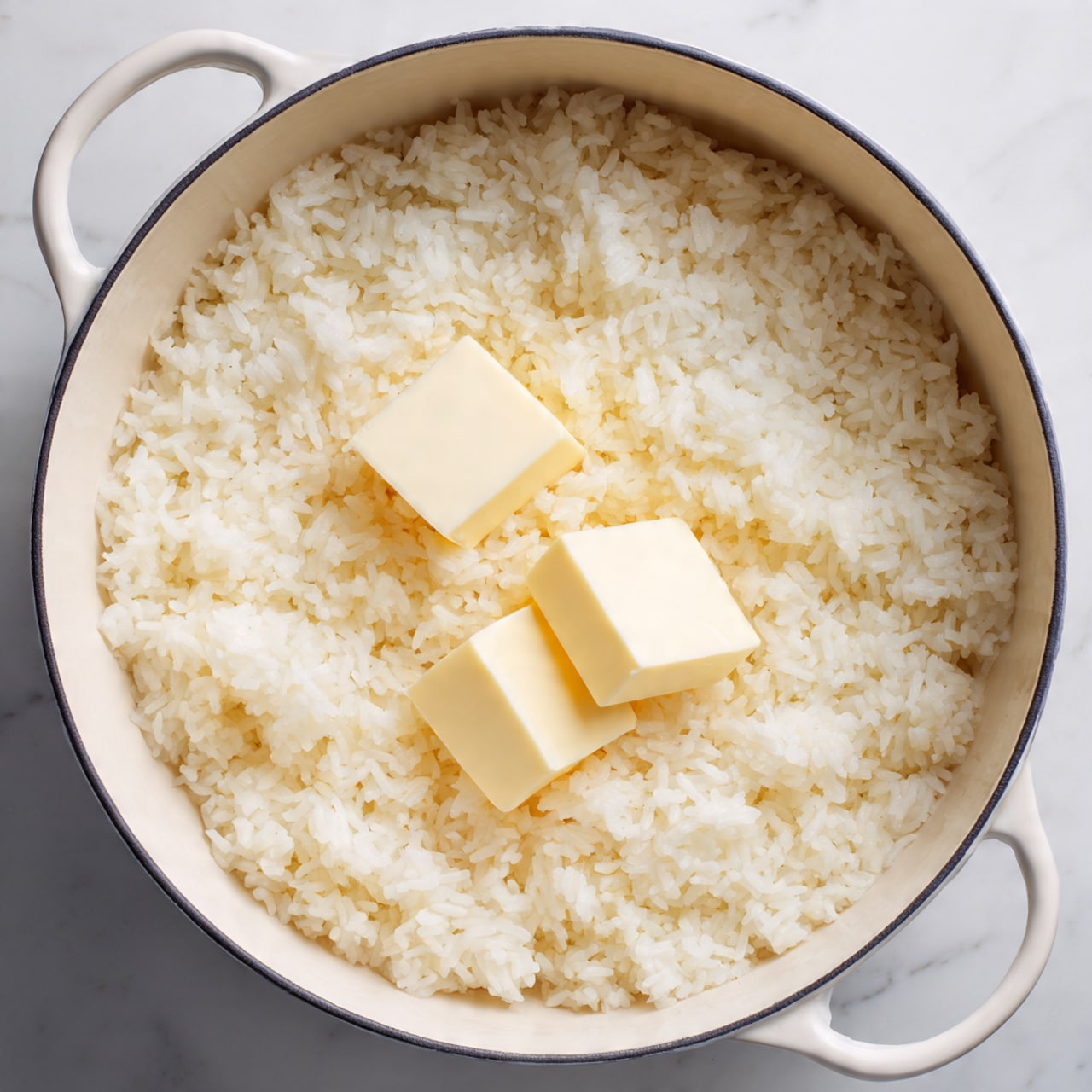A white pot filled with one even layer of cooked white rice that looks fluffy and soft, with two small square pats of pale yellow butter placed near the center on top of the rice. The pot is set on a white marbled surface, and the overall scene looks clean and simple with soft natural light. photo taken with an iphone --ar 4:5 --v 7