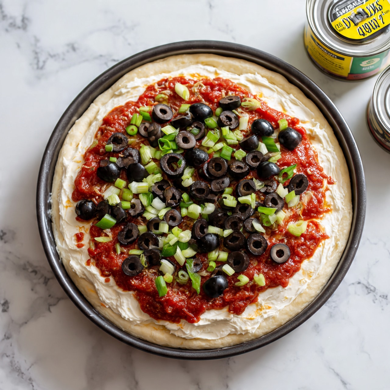A clear glass round dish on a white marbled surface with three visible layers: the bottom layer is white and creamy, the middle layer is red sauce, and the top layer is a mix of black olive slices, small green pieces, and light pink chopped shrimp scattered evenly over the surface. In the background on the same white marbled surface, there is a clear glass bowl filled with several whole cooked shrimp that are pink with white sections. Photo taken with an iphone --ar 4:5 --v 7