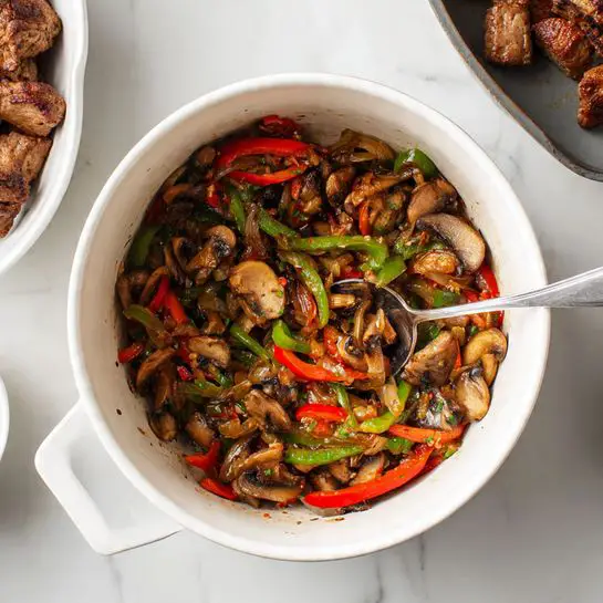 A large white pot filled with a creamy brown soup that has visible red pepper strips and bits of onion mixed in, topped with a pile of shredded white cheese in the center. A dark green ladle is resting in the pot, partially covered by the cheese. To the left, there is a white bowl with thin slices of cooked beef. On the right, a metal measuring cup holds more shredded white cheese. All items are set on a white marbled surface. Photo taken with an iphone --ar 4:5 --v 7